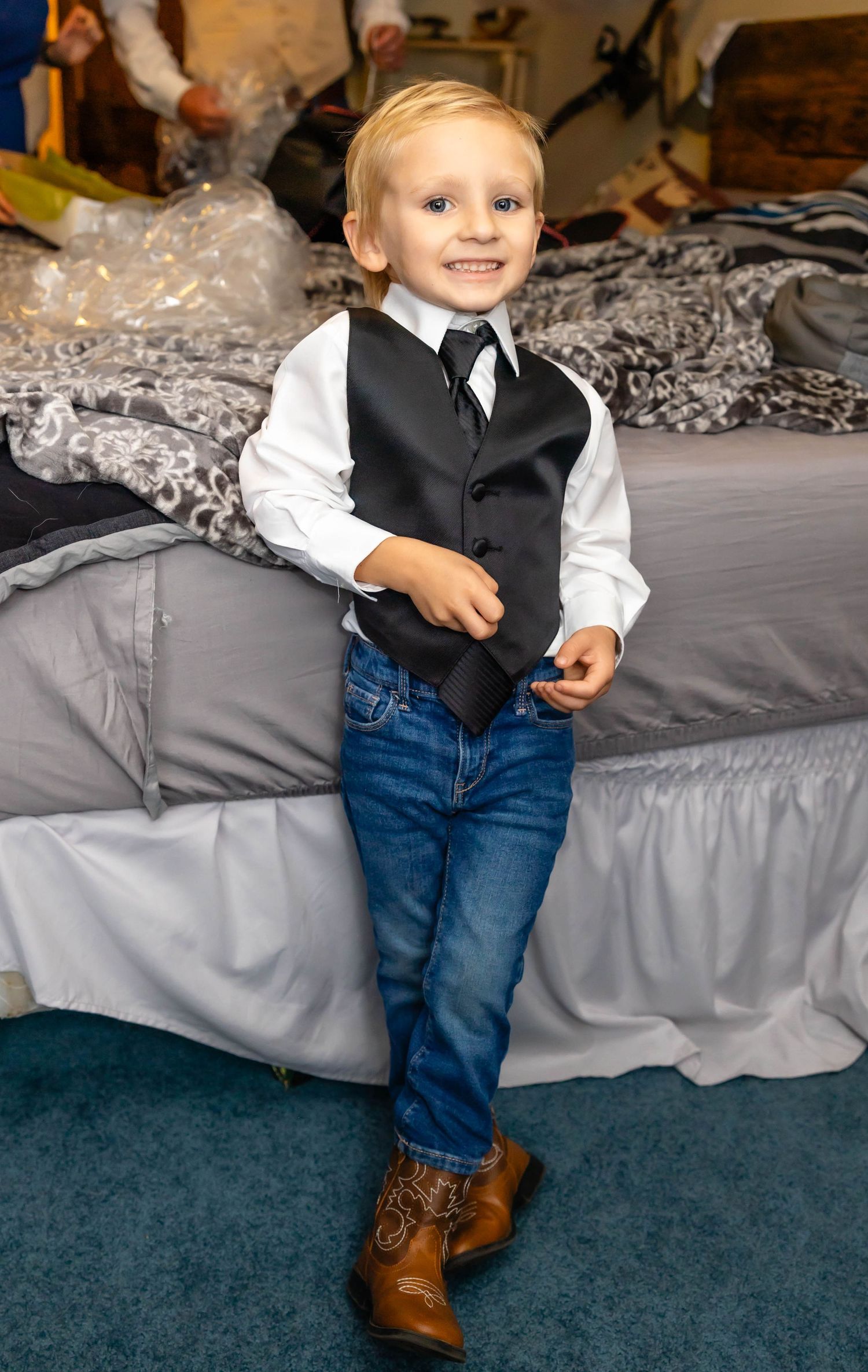 A young ring bearer in a black vest and jeans poses with a bright smile.