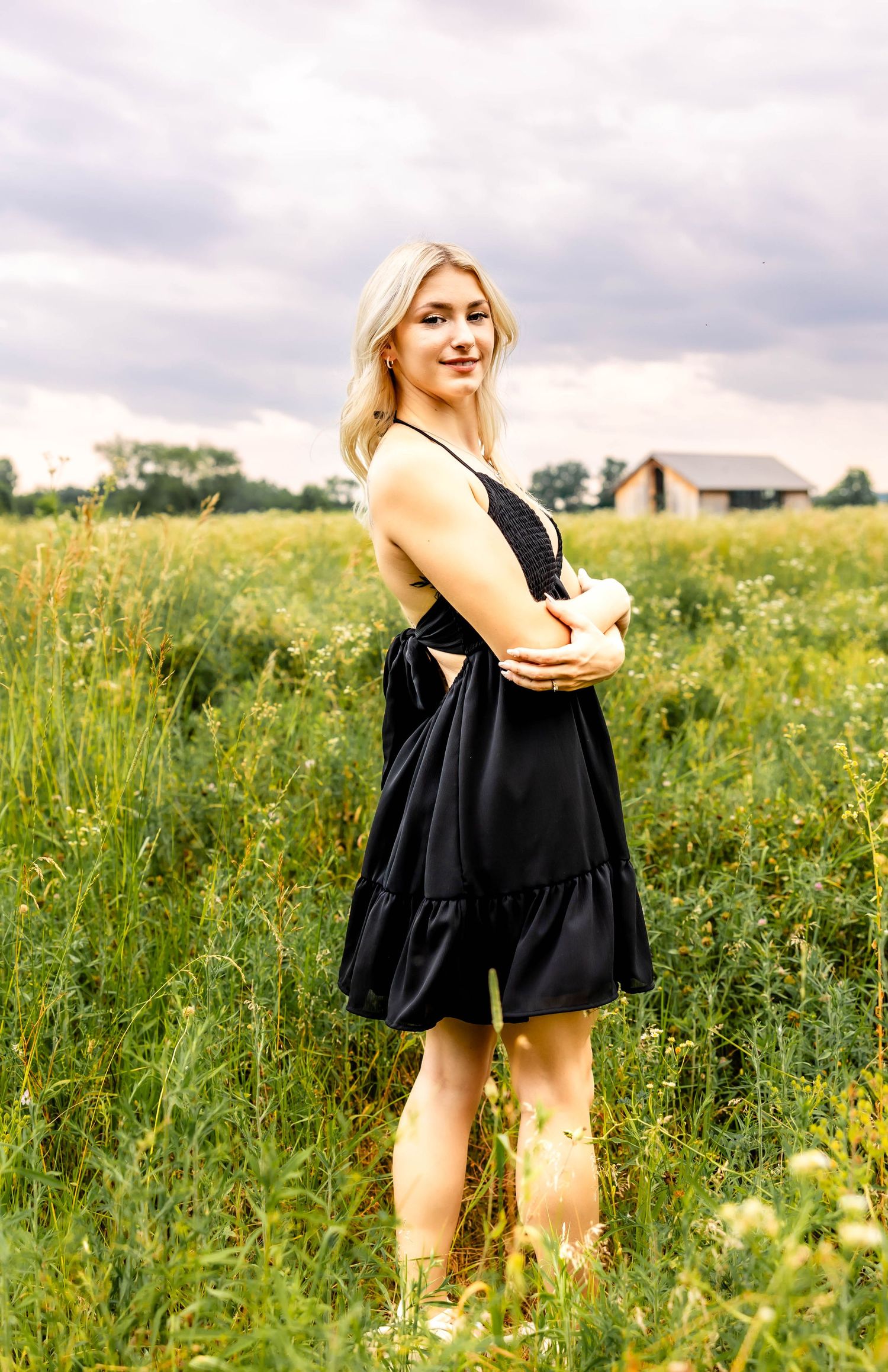 Summer portrait in a black dress standing among wildflowers with a farmhouse in background.