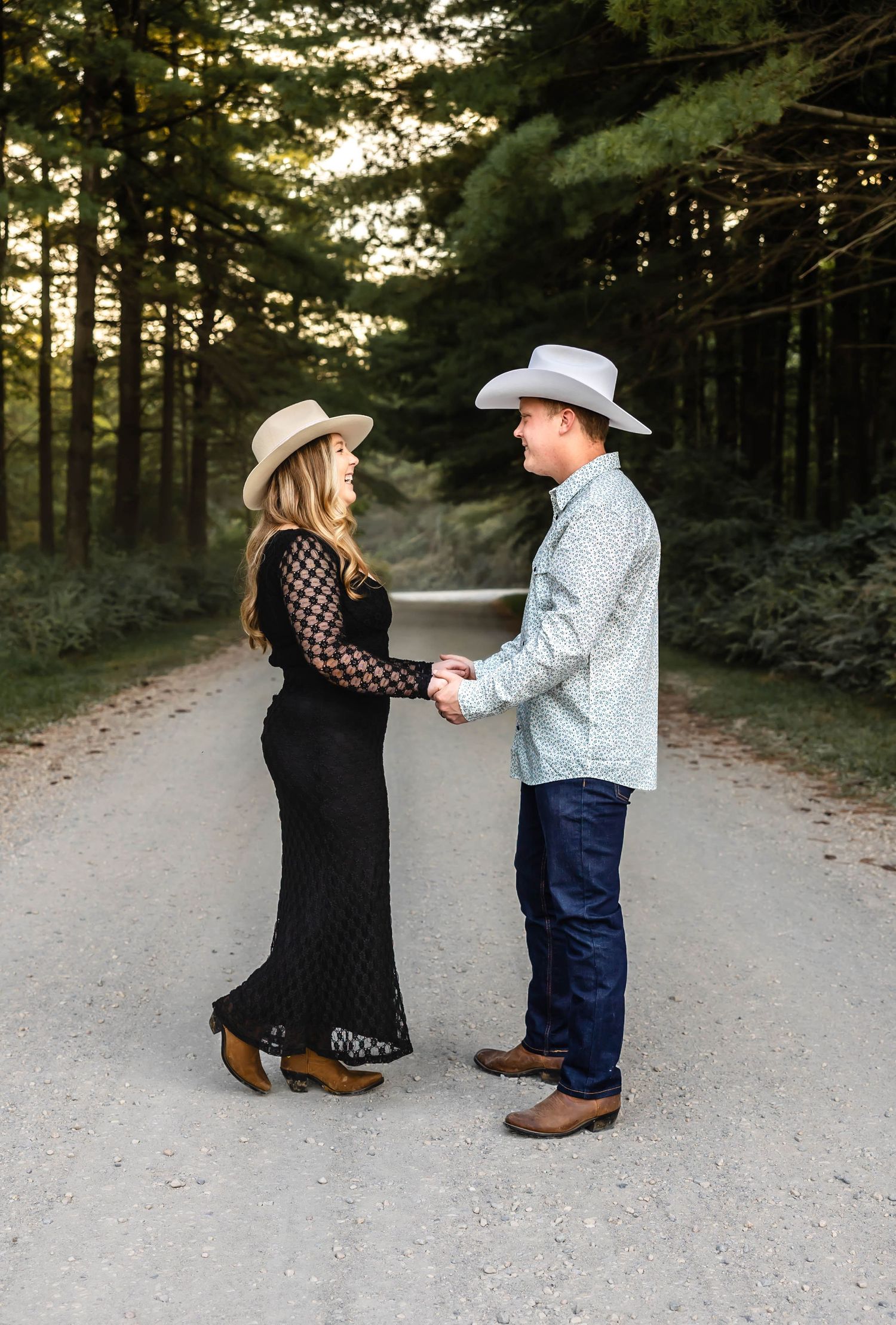 Couple in western attire hold hands while standing on forest path during engagement session.