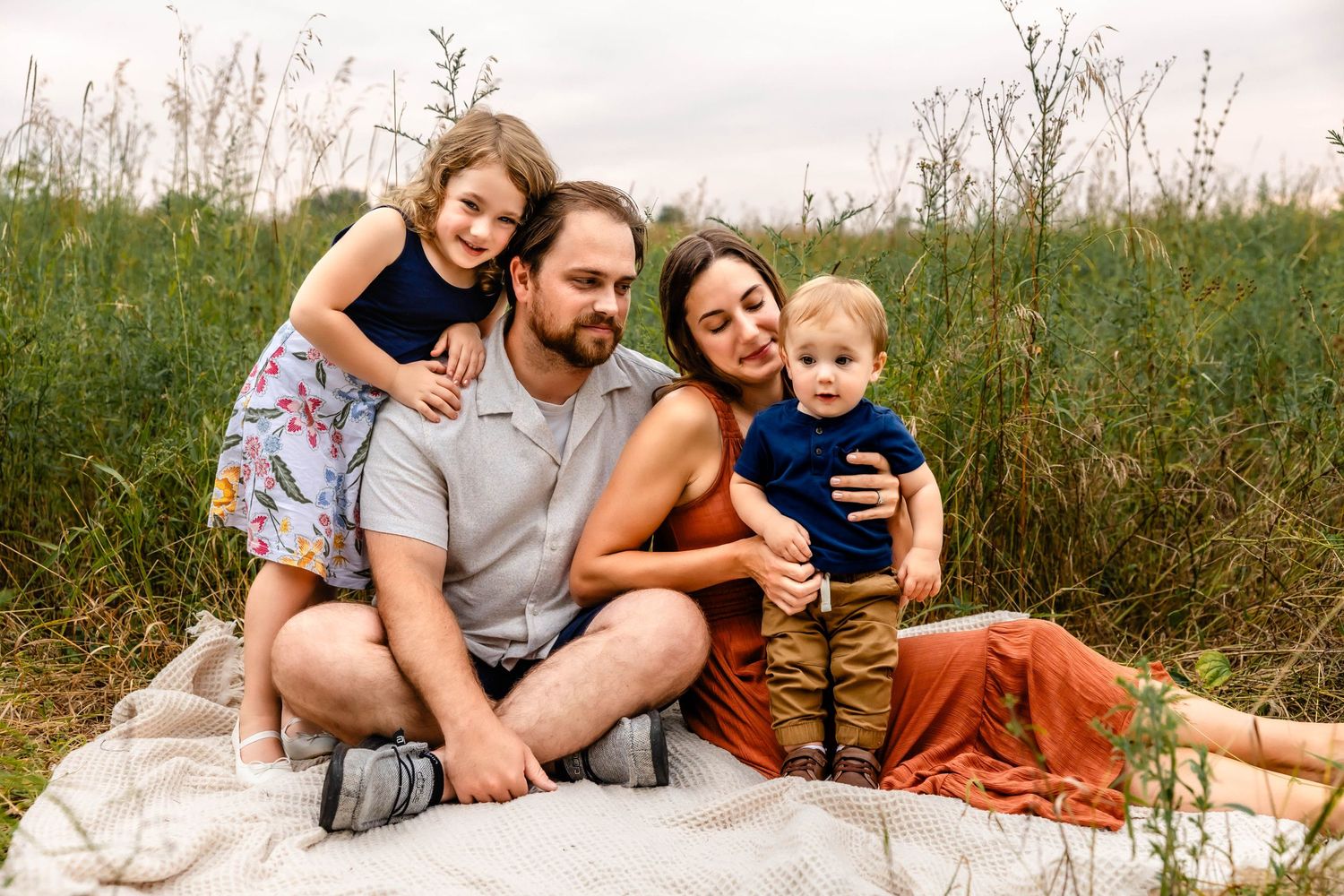 A happy family shares a tender moment together while sitting on a blanket in a meadow on a cloudy day.