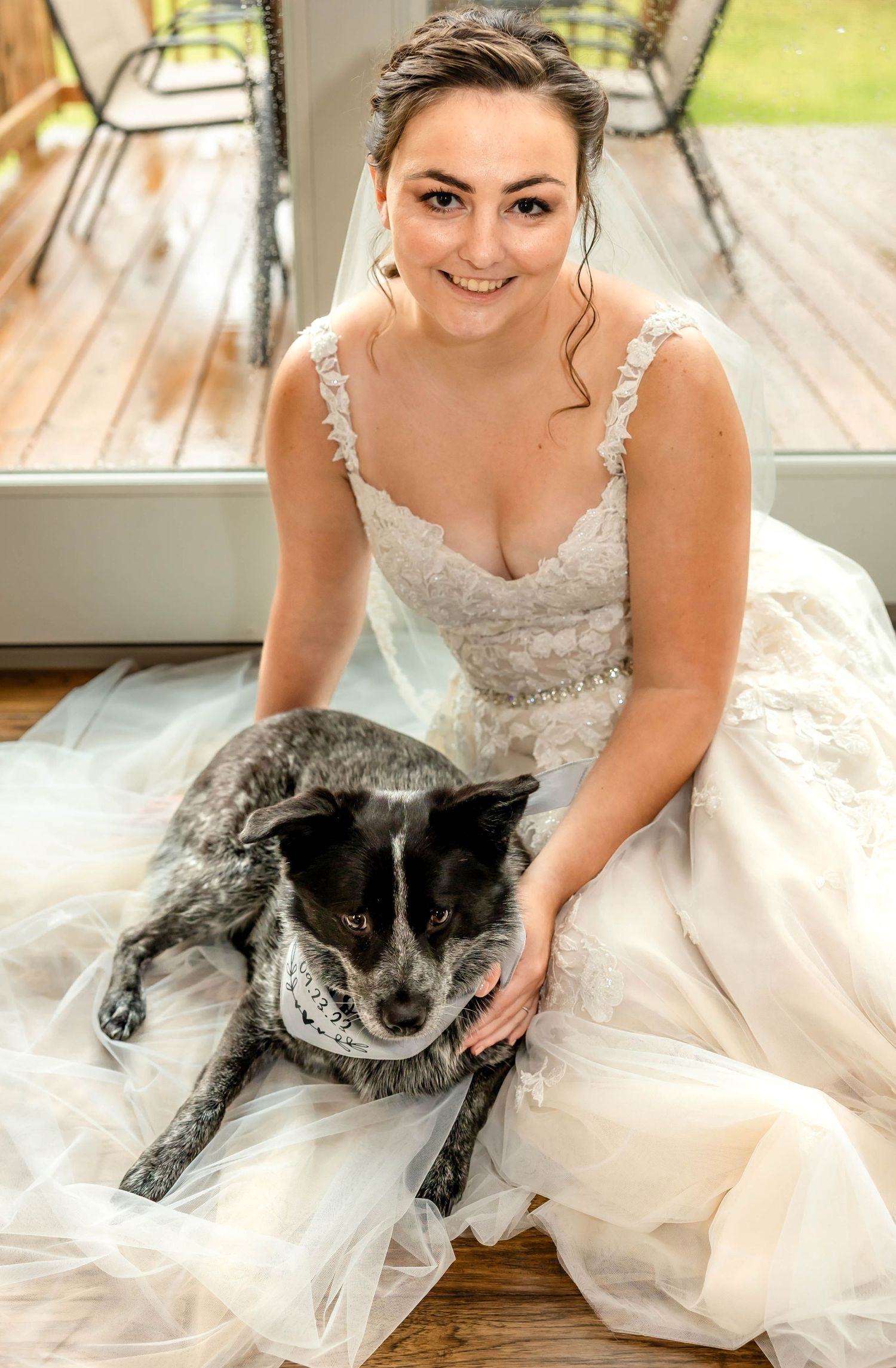 A bride in a lace wedding dress poses with her gray and black dog on a deck.