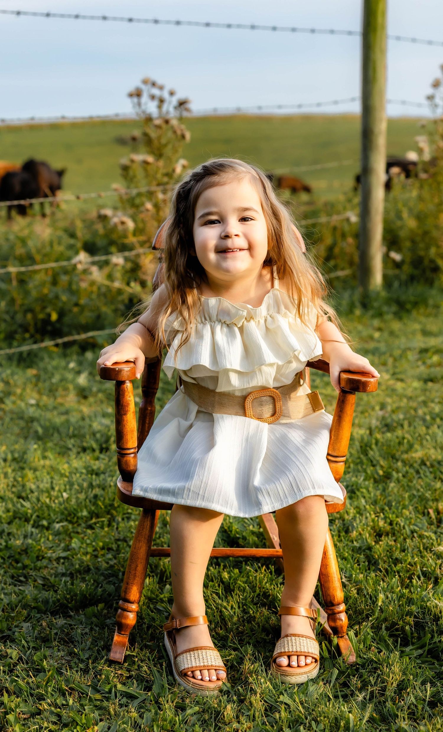 Candid outdoor portrait on a wooden chair with countryside and grazing cattle in background.