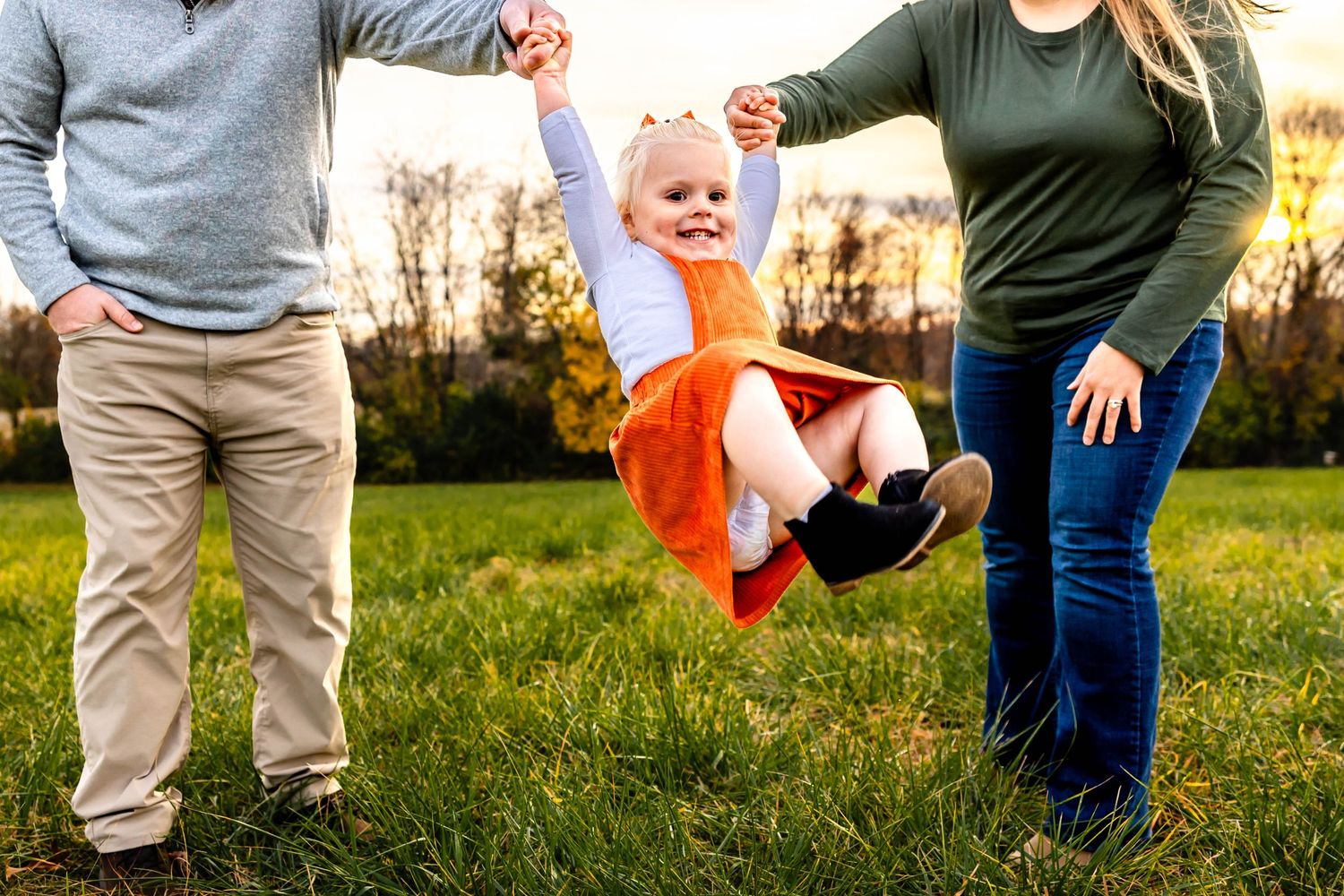 Parents swinging child wearing orange jumper dress in outdoor field at sunset.