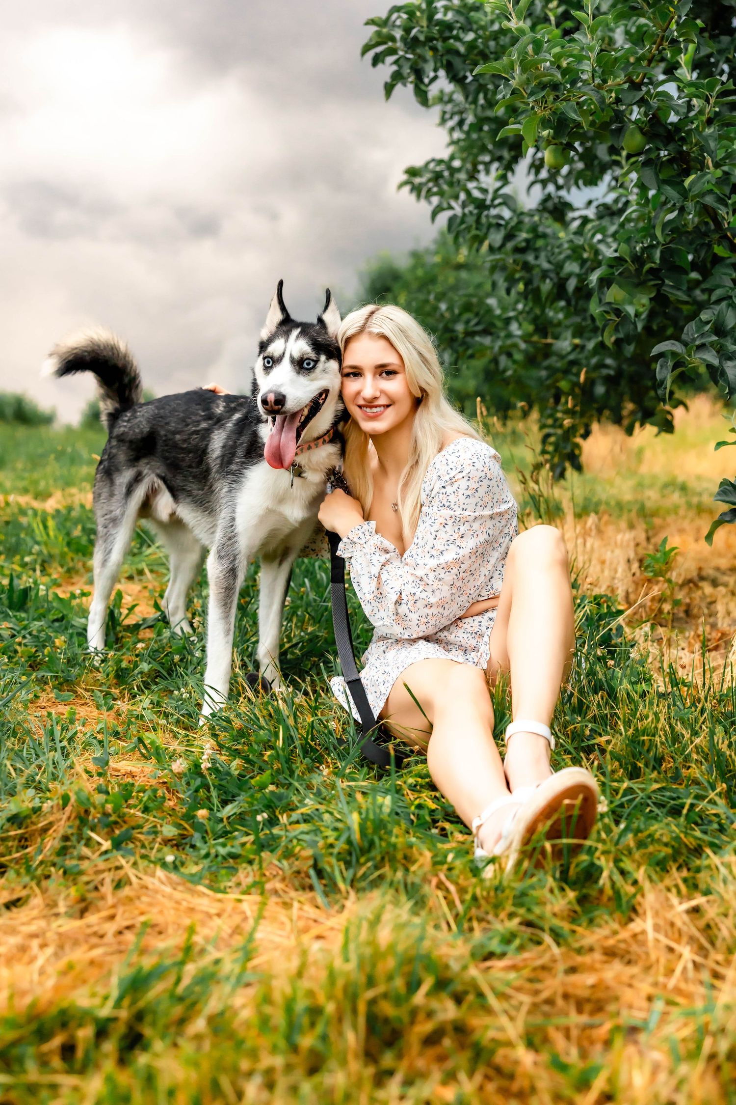Woman in lace dress poses with Husky dog in grassy field during sunset photoshoot.
