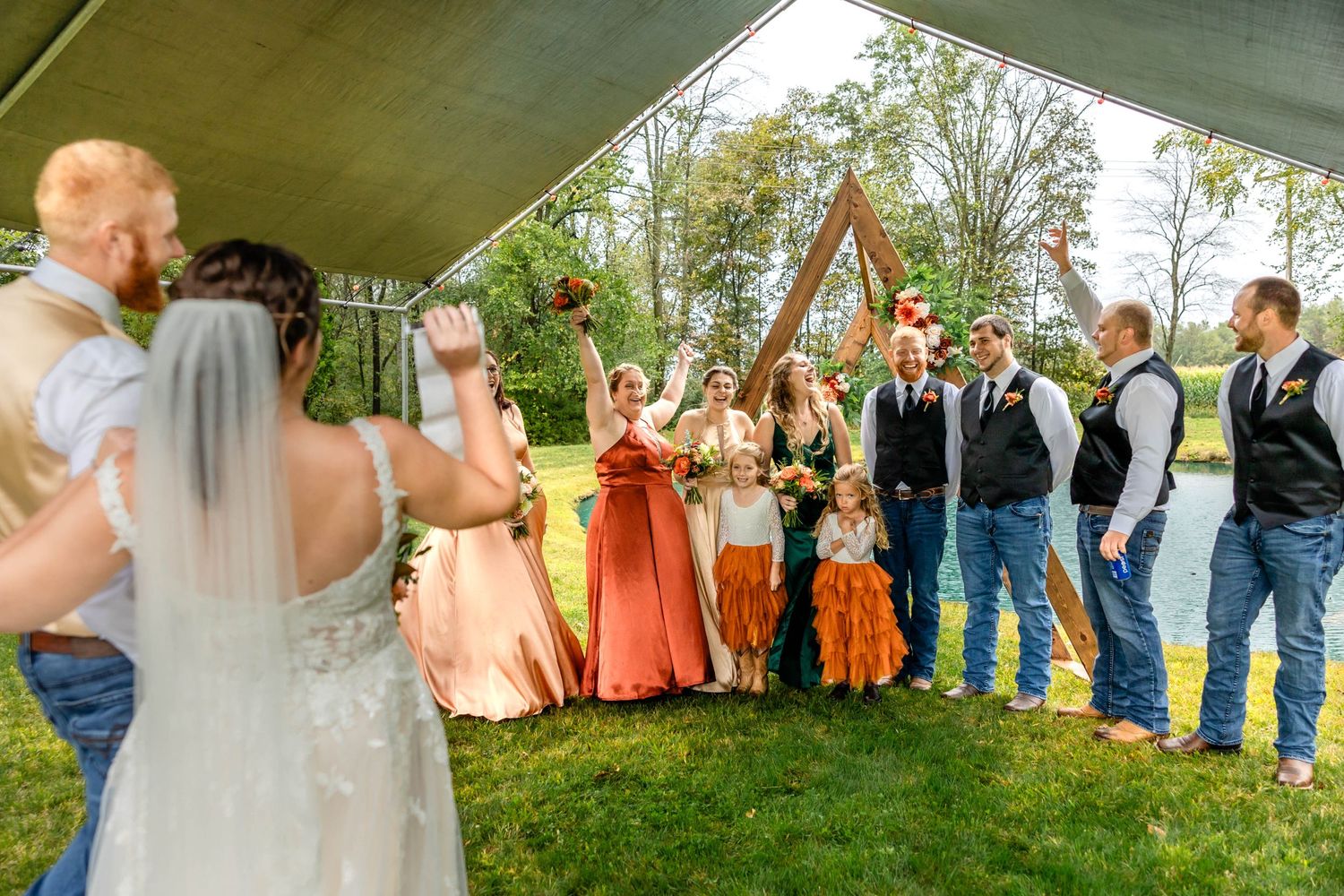 Wedding party poses outdoors under a tent canopy with bridesmaids in coral dresses and groomsmen in casual attire.