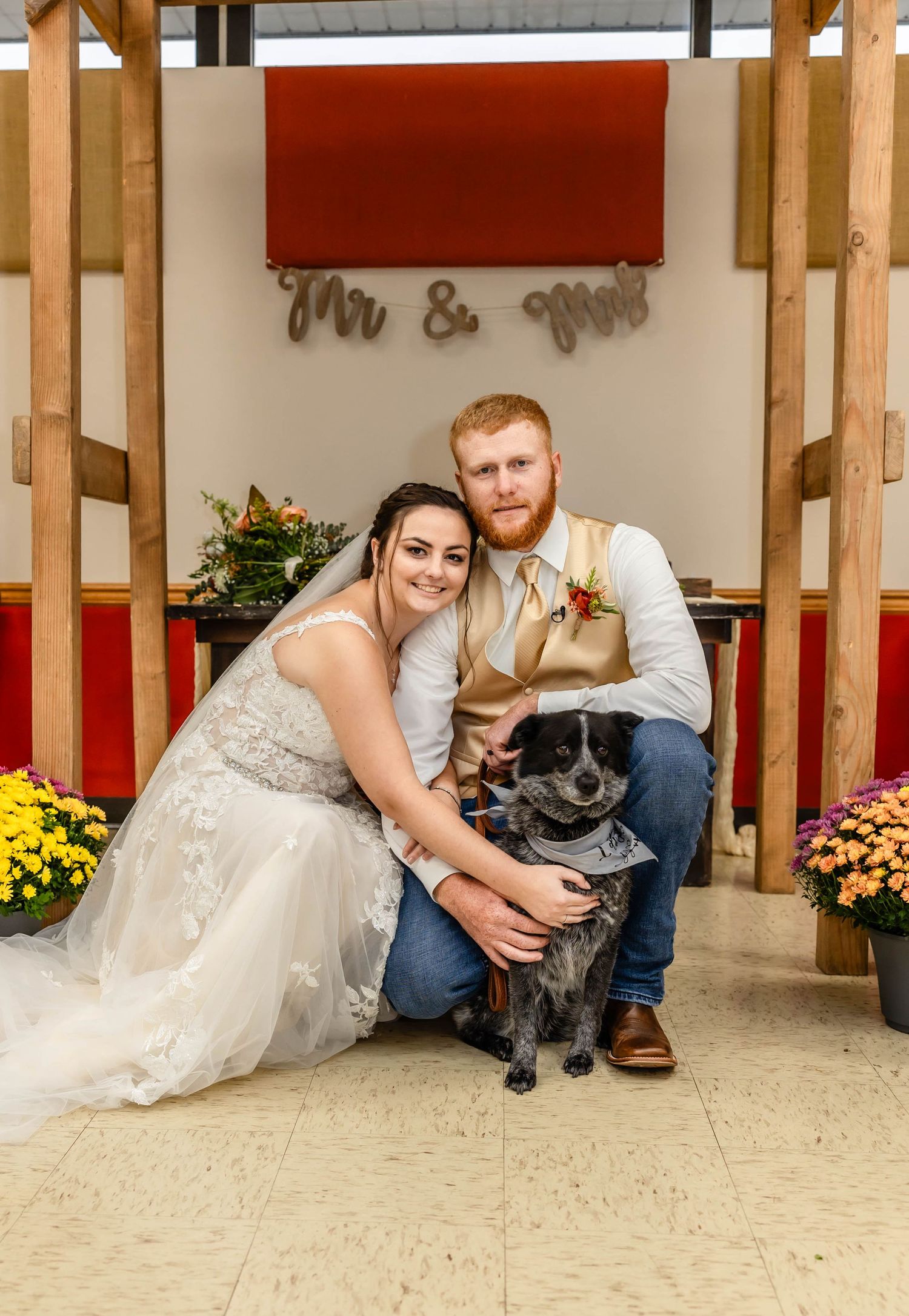 Newlyweds pose with their black dog under a rustic wooden arch with yellow mums.