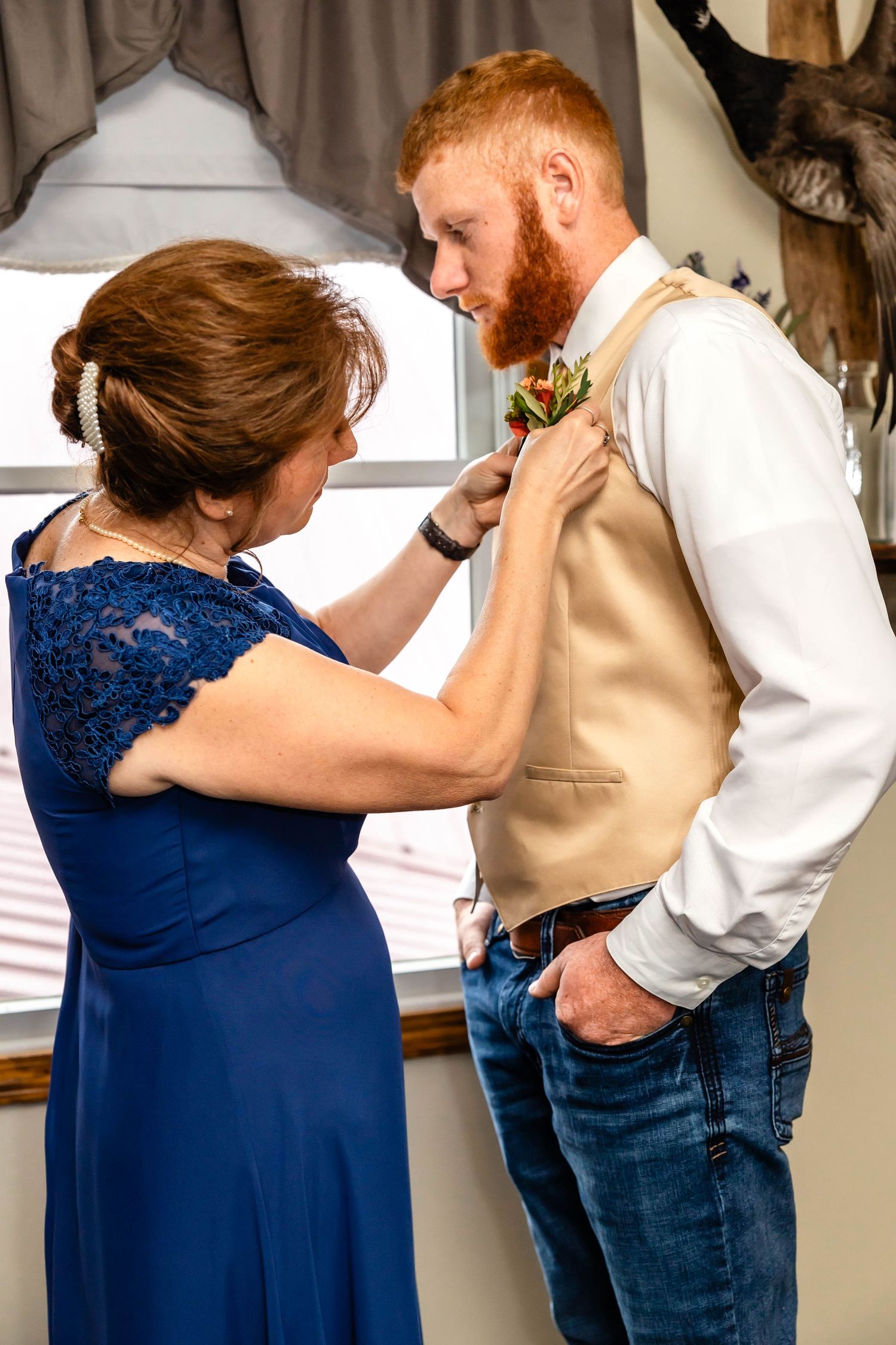 A woman in blue adjusts a boutonniere on someone wearing a tan vest.