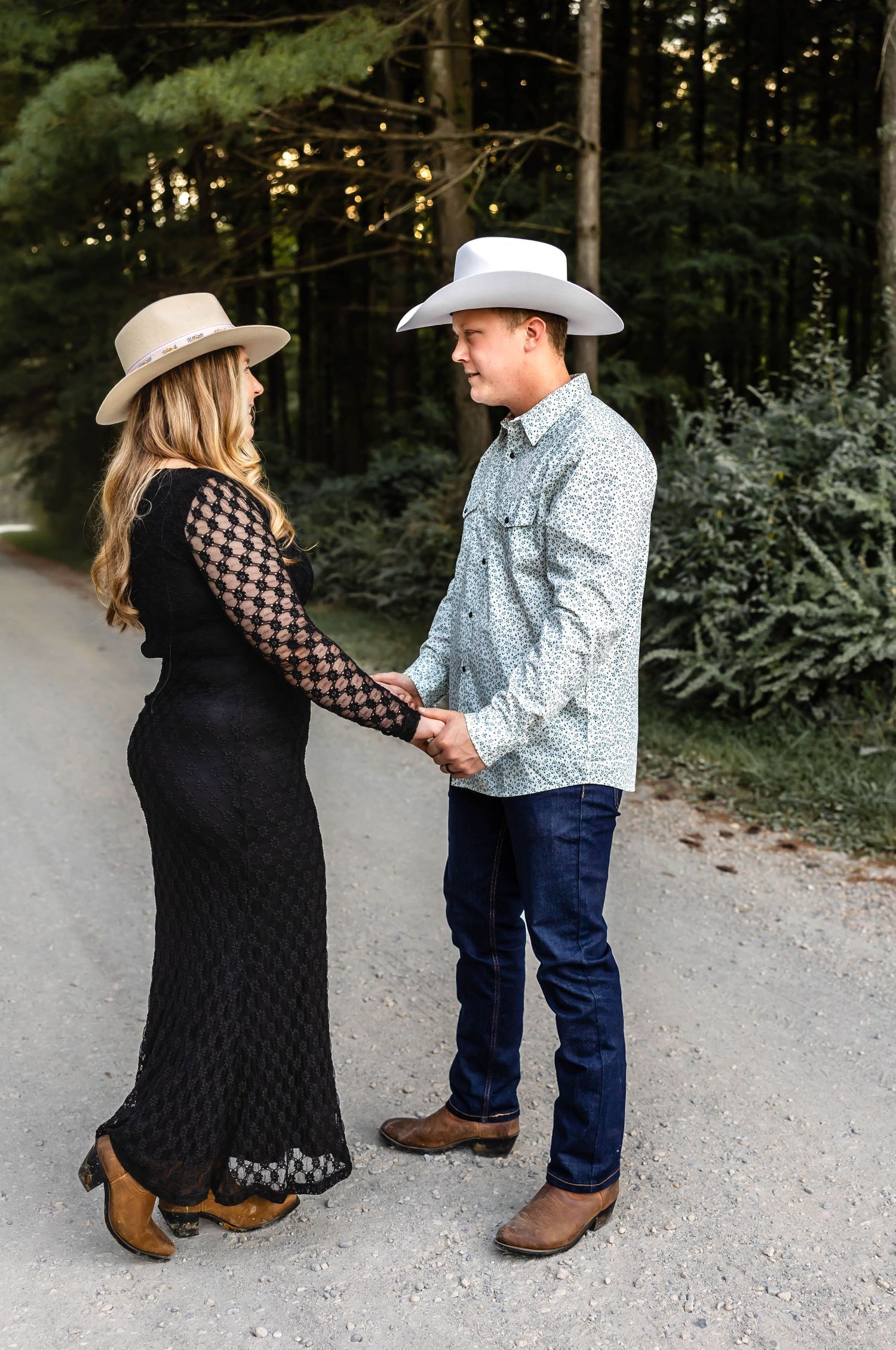A couple in western attire holding hands on a wooded path, wearing cowboy hats and country-inspired outfits.