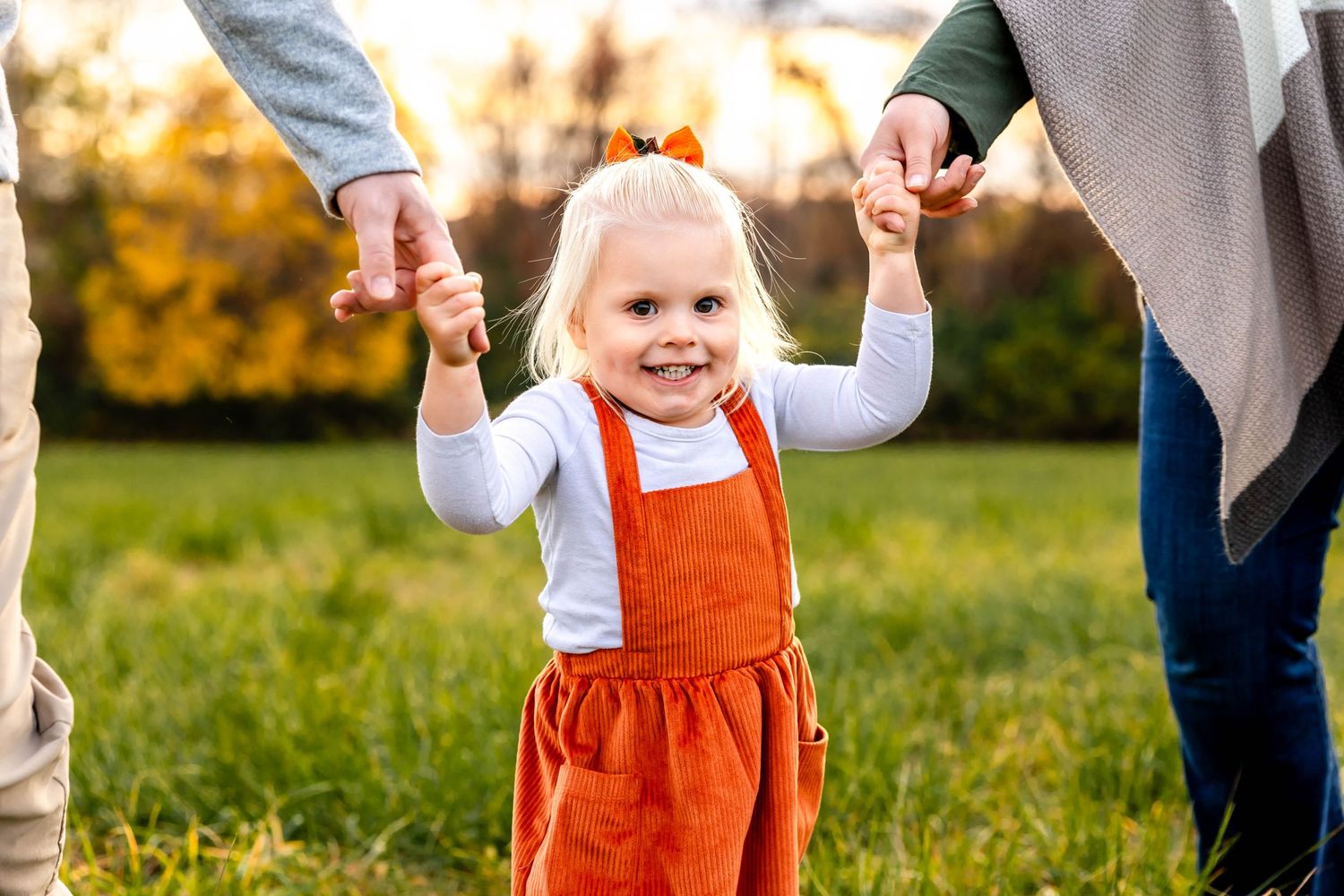 A small child in an orange jumper dress holding hands with adults while walking outdoors during autumn.