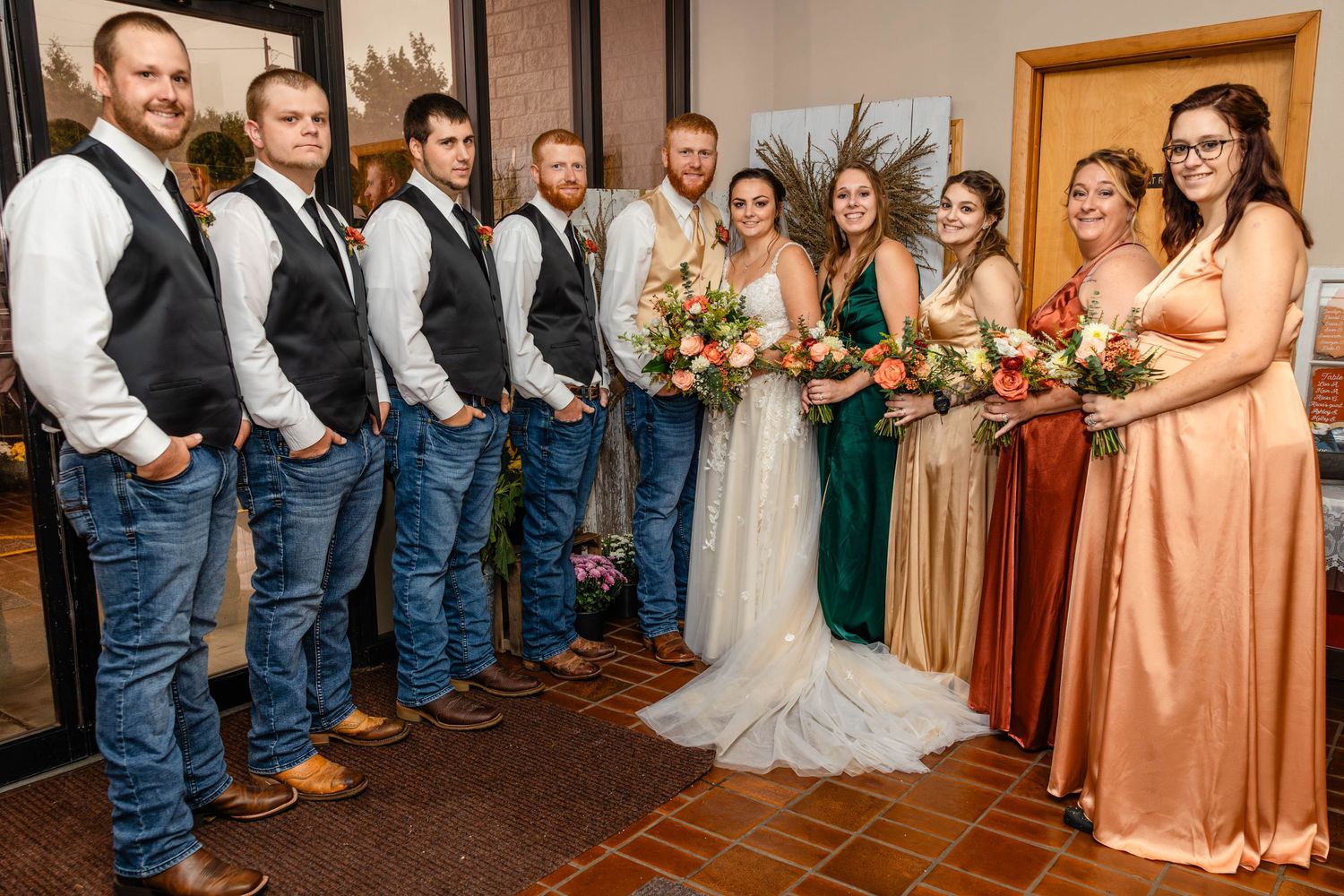Wedding party in blue jeans, vests and autumn-colored dresses poses on wooden floor.