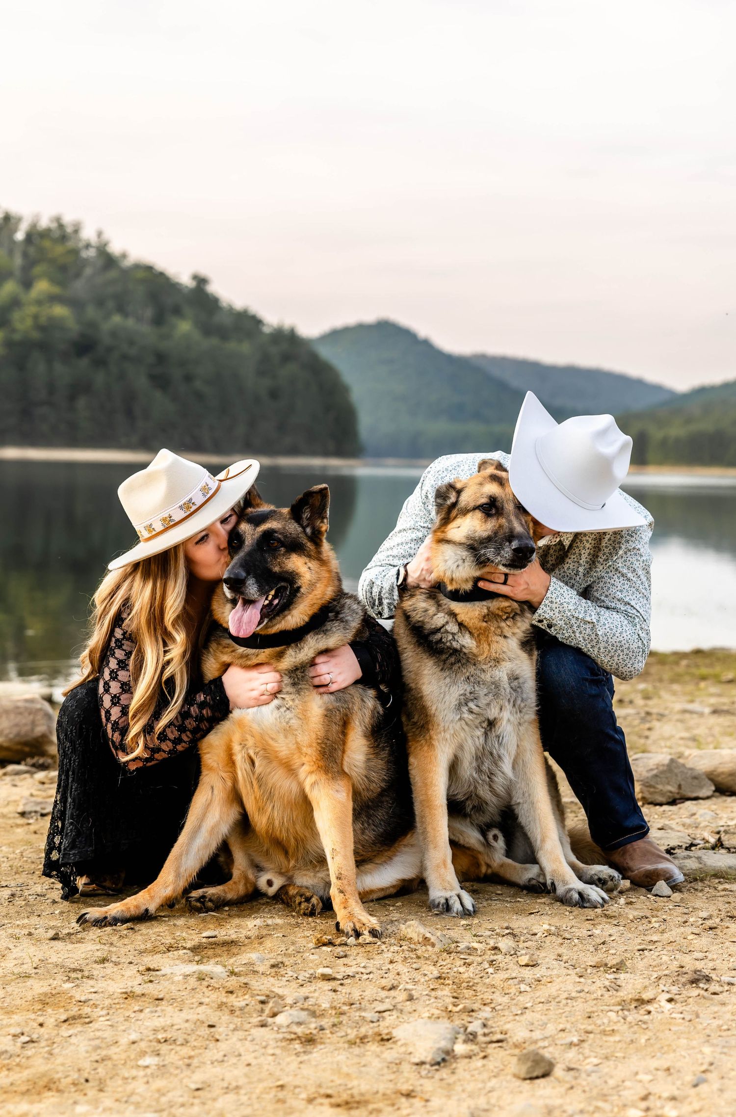 A couple in western attire cuddles with their German Shepherd dogs on a lakeside shore.