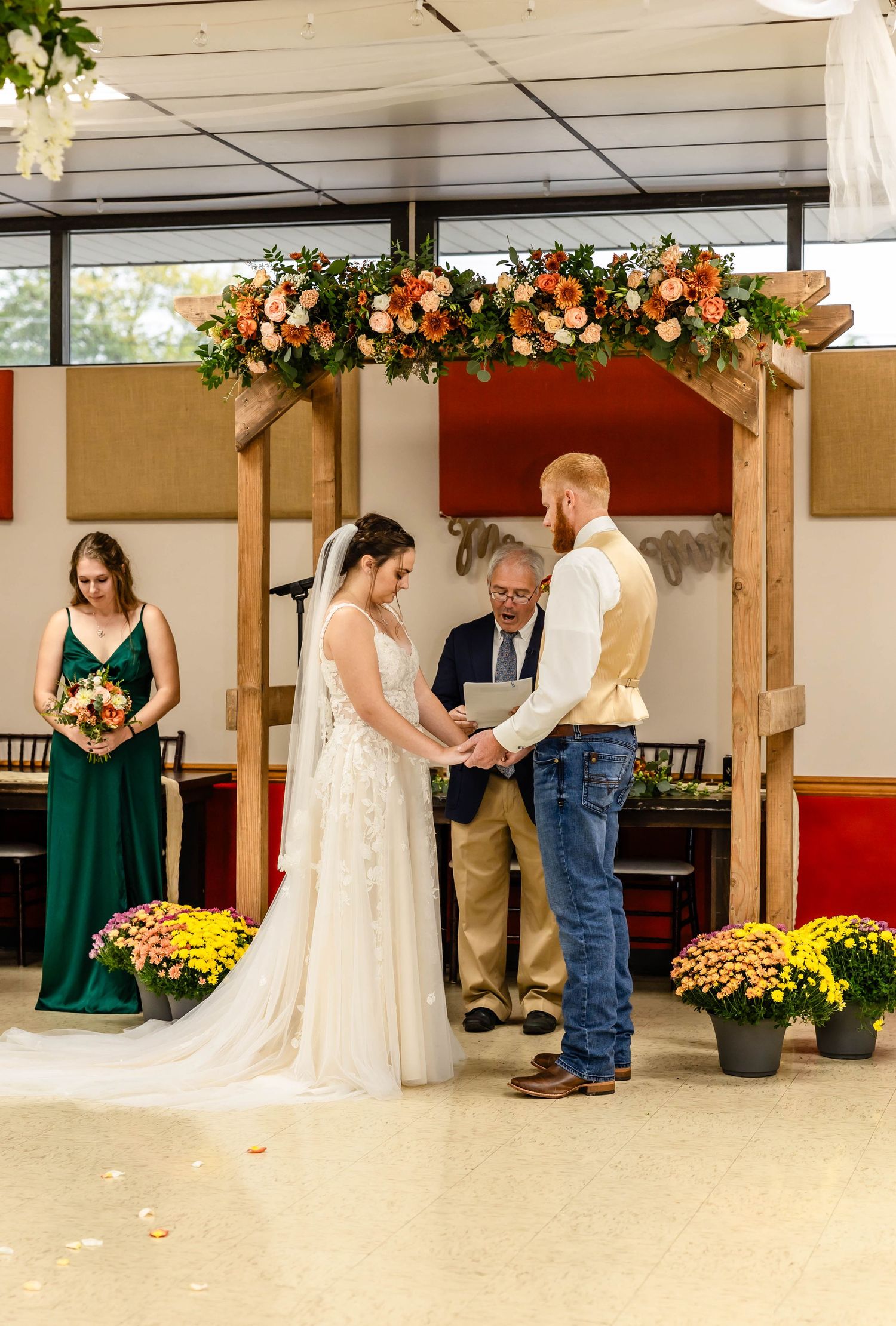 A wedding ceremony under a wooden arch decorated with orange and peach flowers and potted mums.