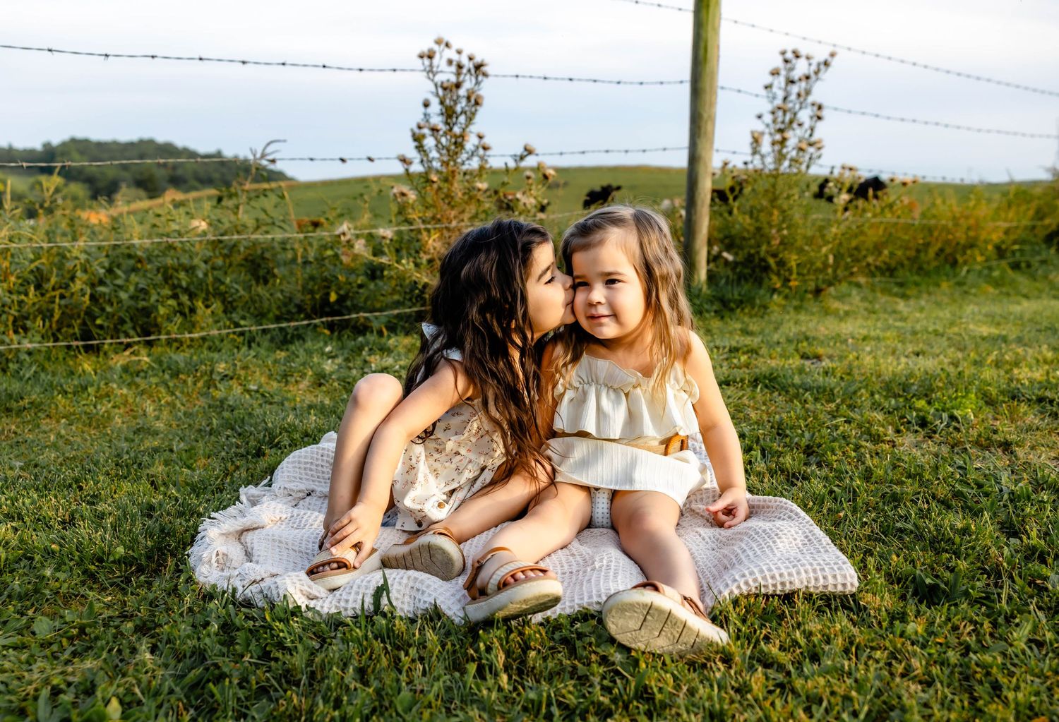 Two children in light dresses sitting close together on blanket with scenic countryside backdrop.