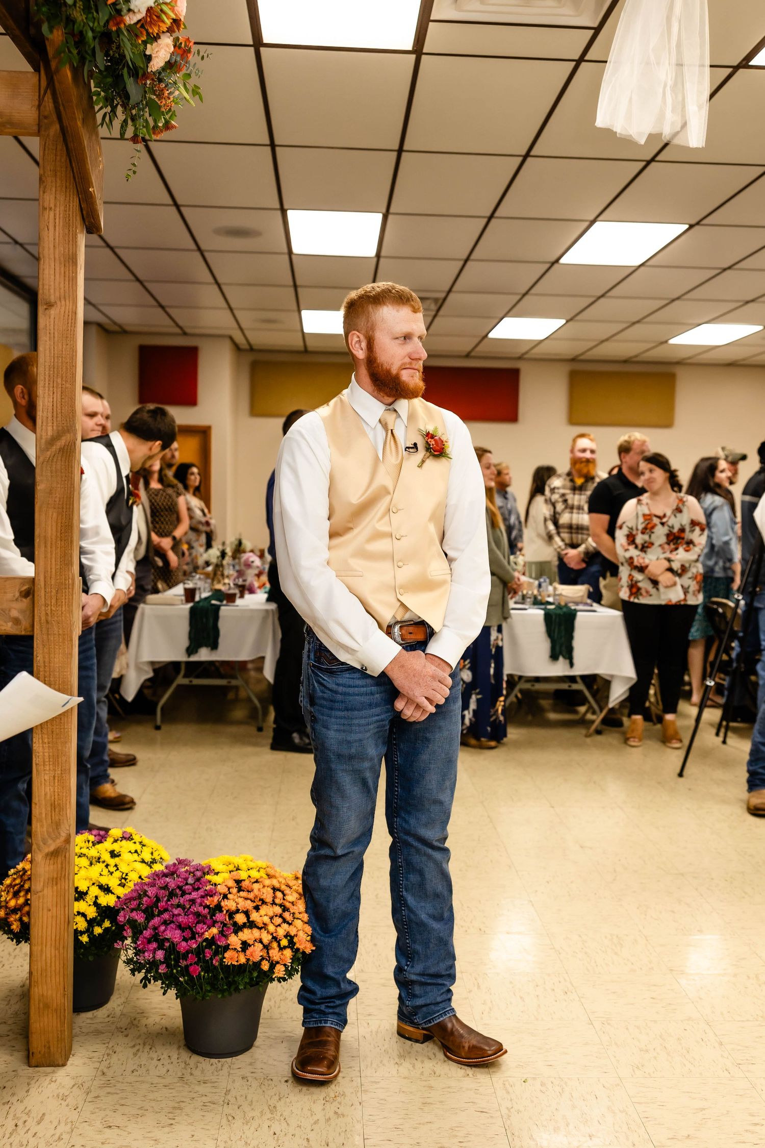 A groom in a tan vest and jeans stands beneath wooden beams decorated with flowers at an indoor ceremony.