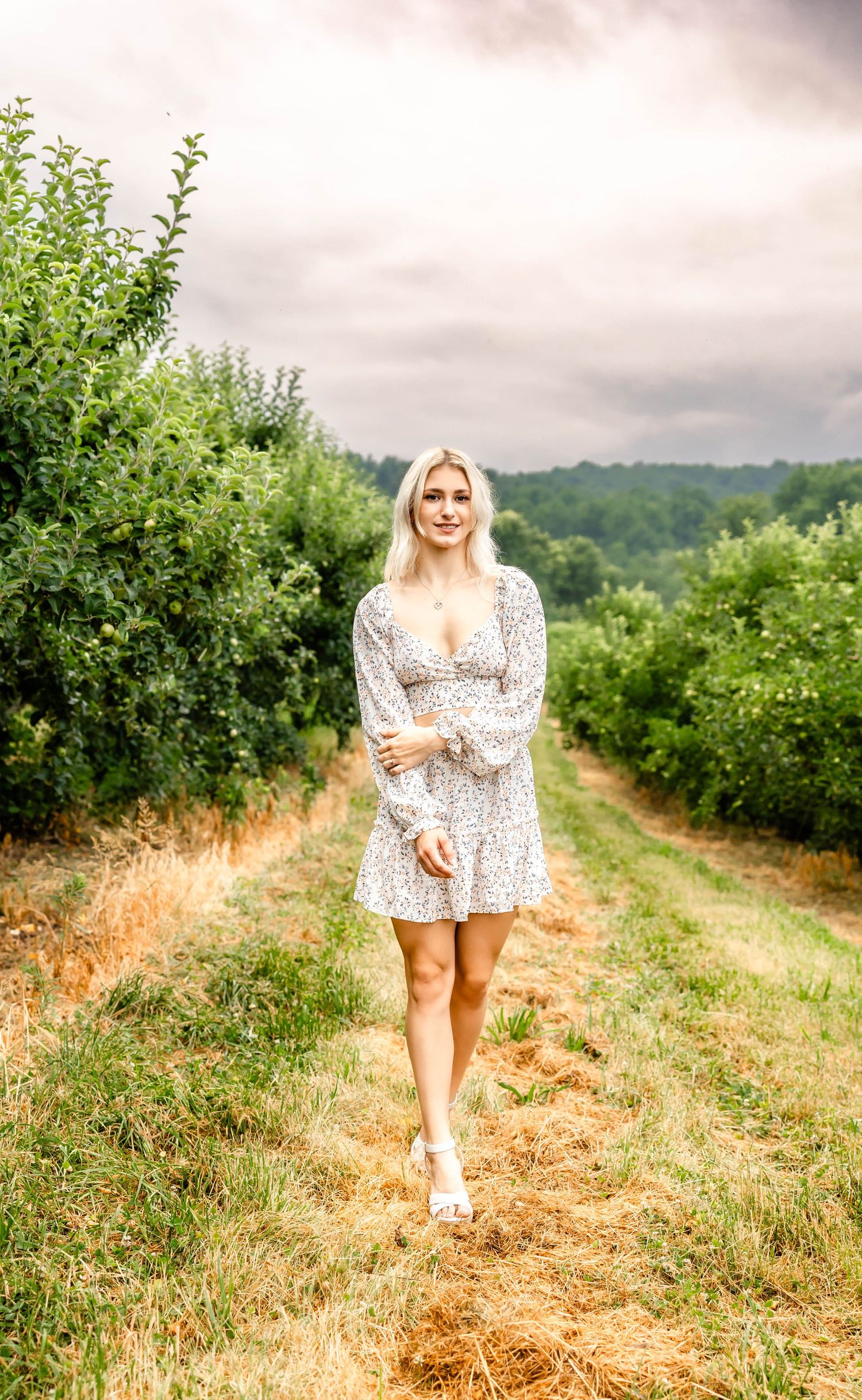 Ethereal outdoor portrait in a white floral dress walking through an orchard path.