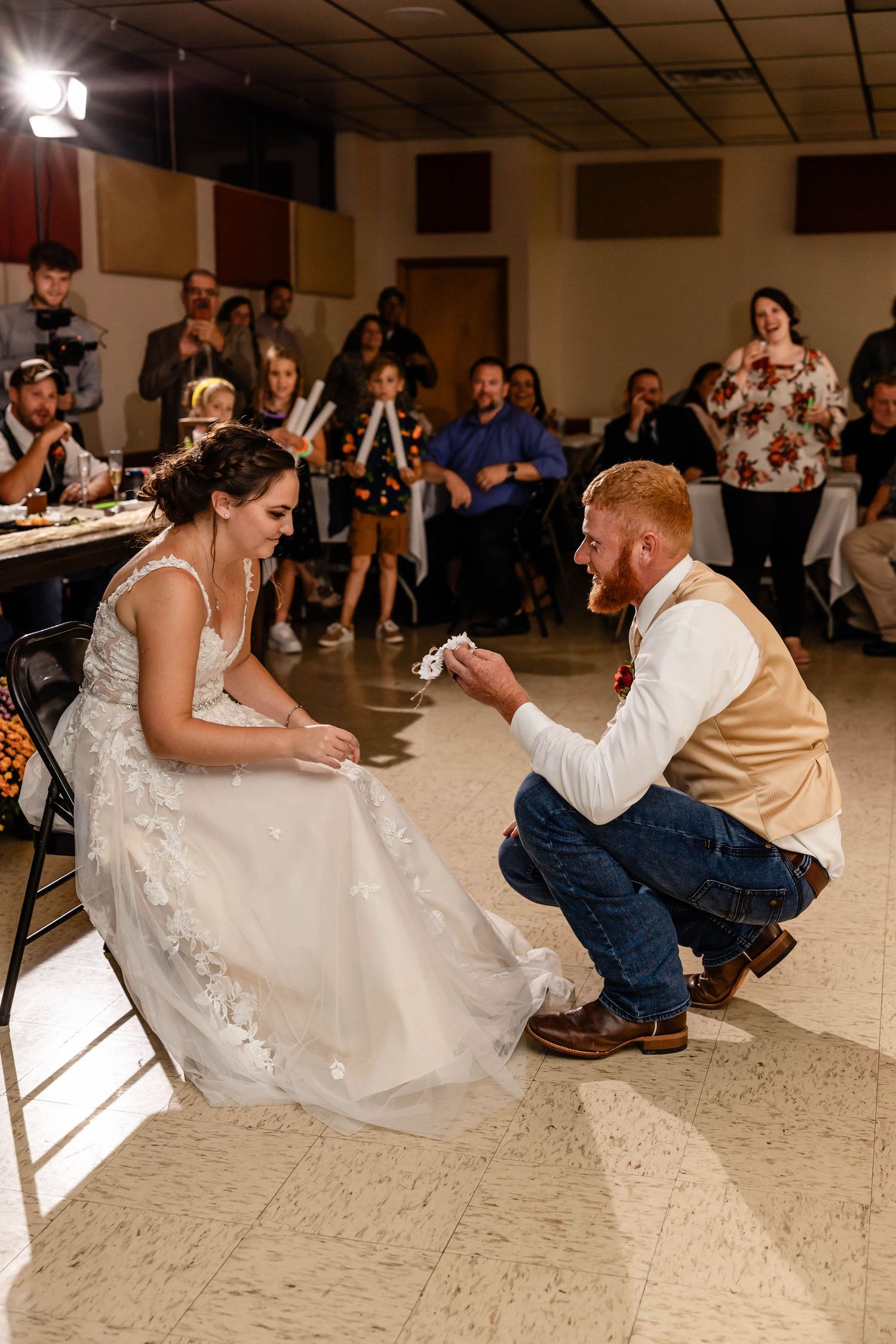 Wedding couple shares playful moment during reception game as guests watch from the background in community hall.