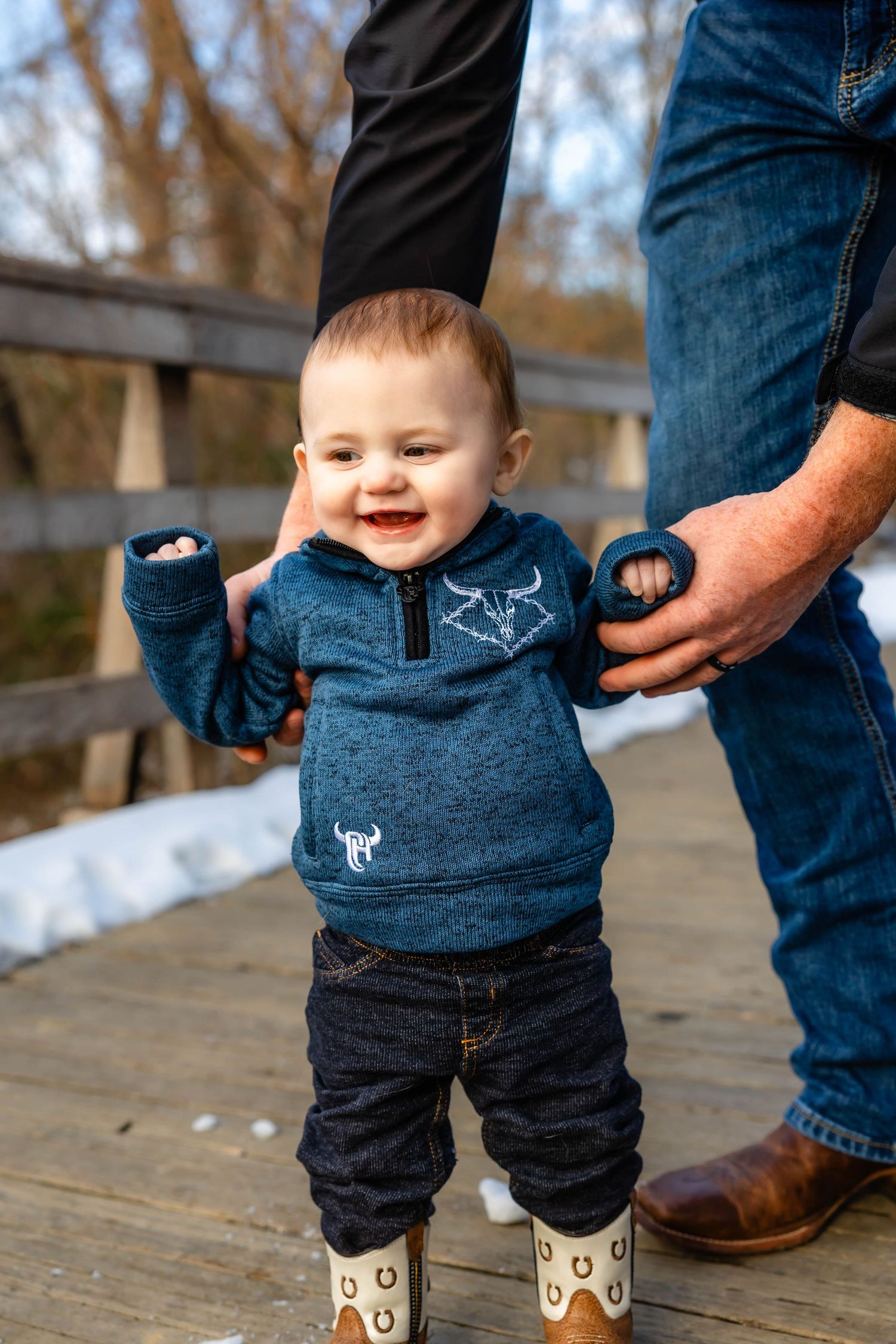 Joyful baby in blue sweater laughing while standing on wooden bridge.
