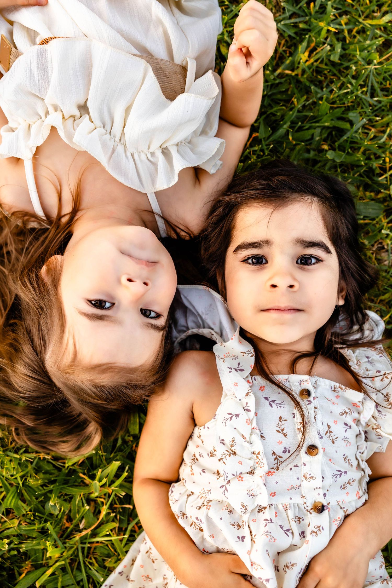 Two people lying on green grass wearing white and floral dresses while looking at the camera.