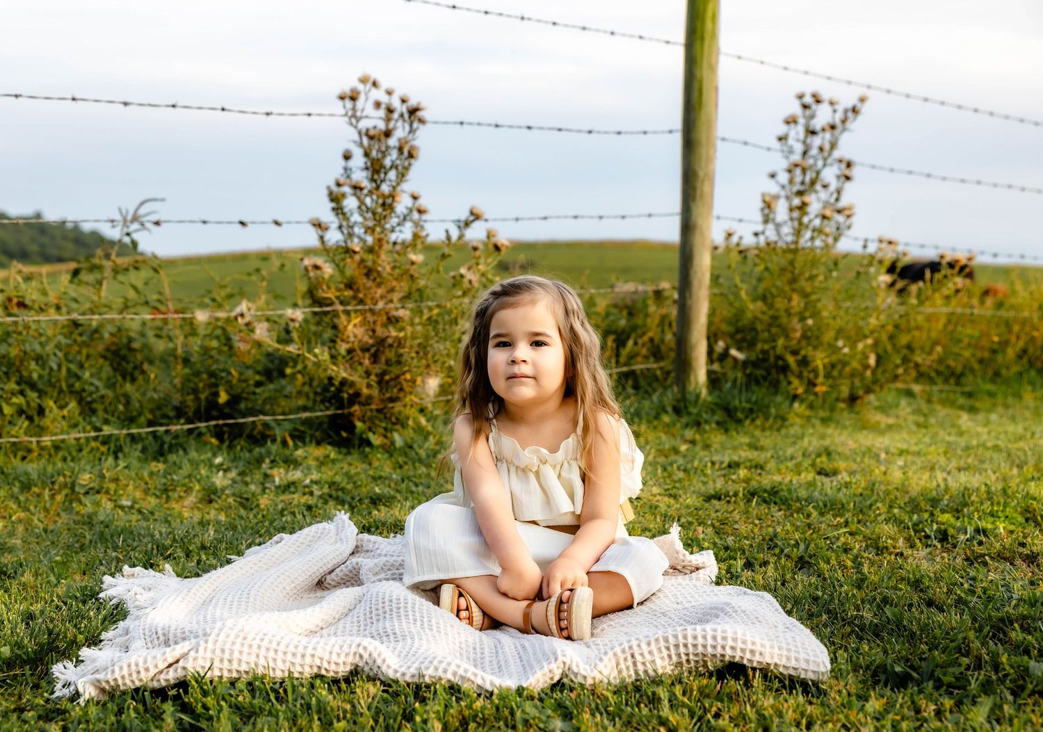 Child sitting cross-legged on white blanket in countryside setting with barbed wire fence and rolling hills behind.