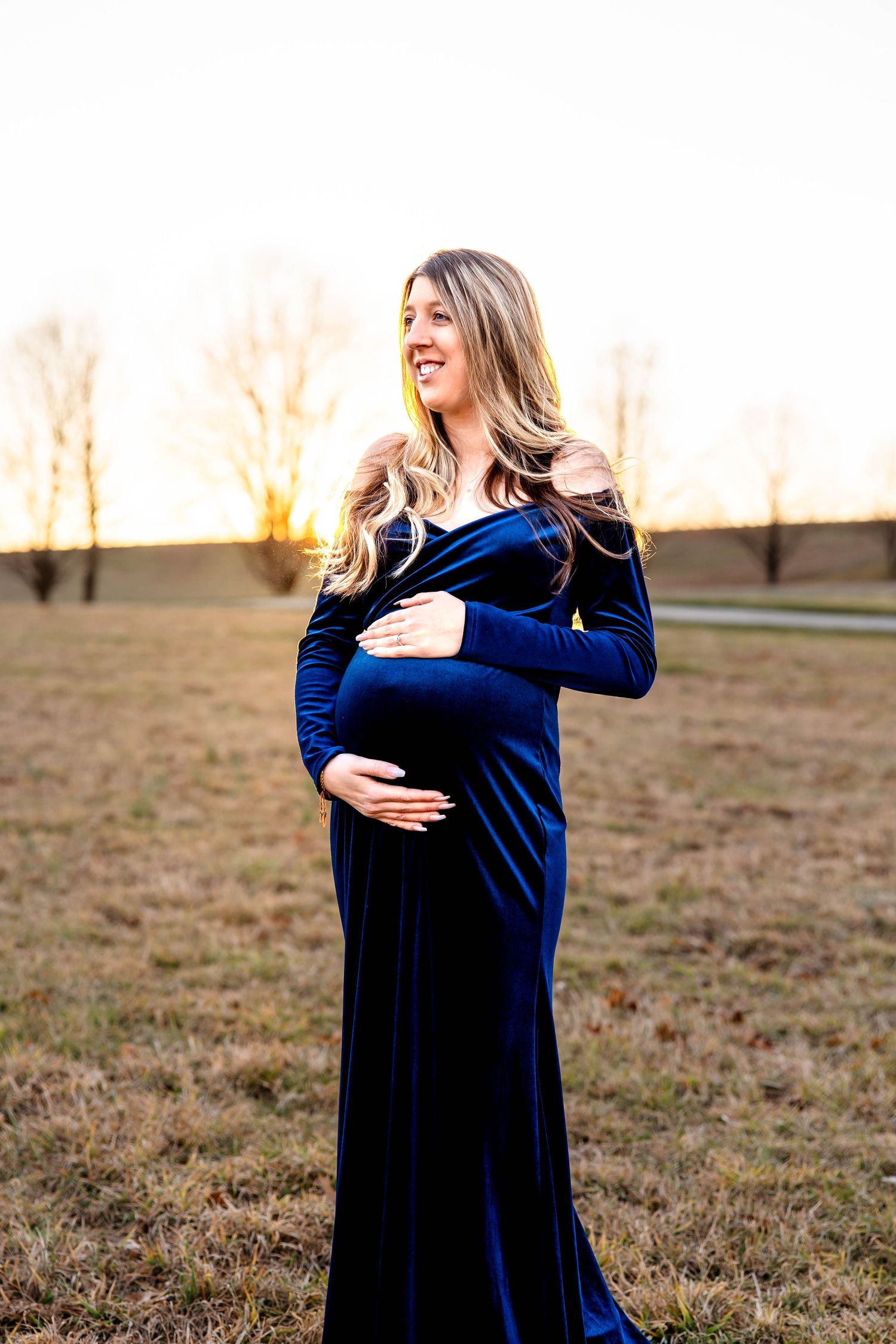 An expectant mother in a royal blue velvet dress poses during sunset in an outdoor maternity session.