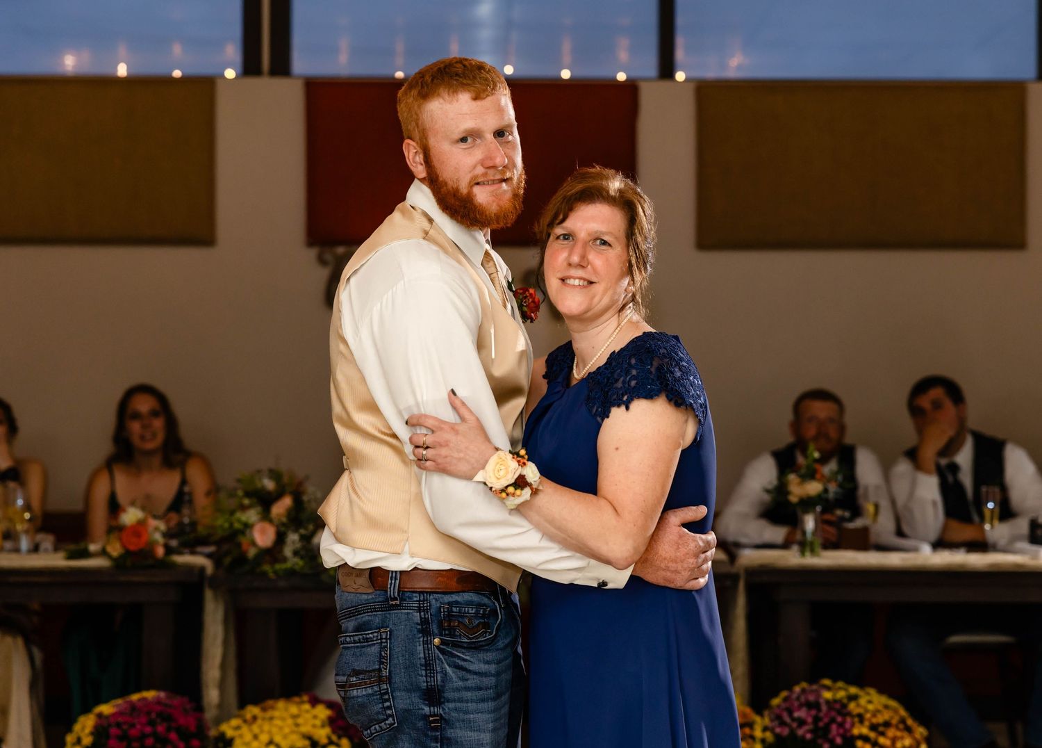 A couple embraces while dancing at their wedding reception with guests and floral decor visible.