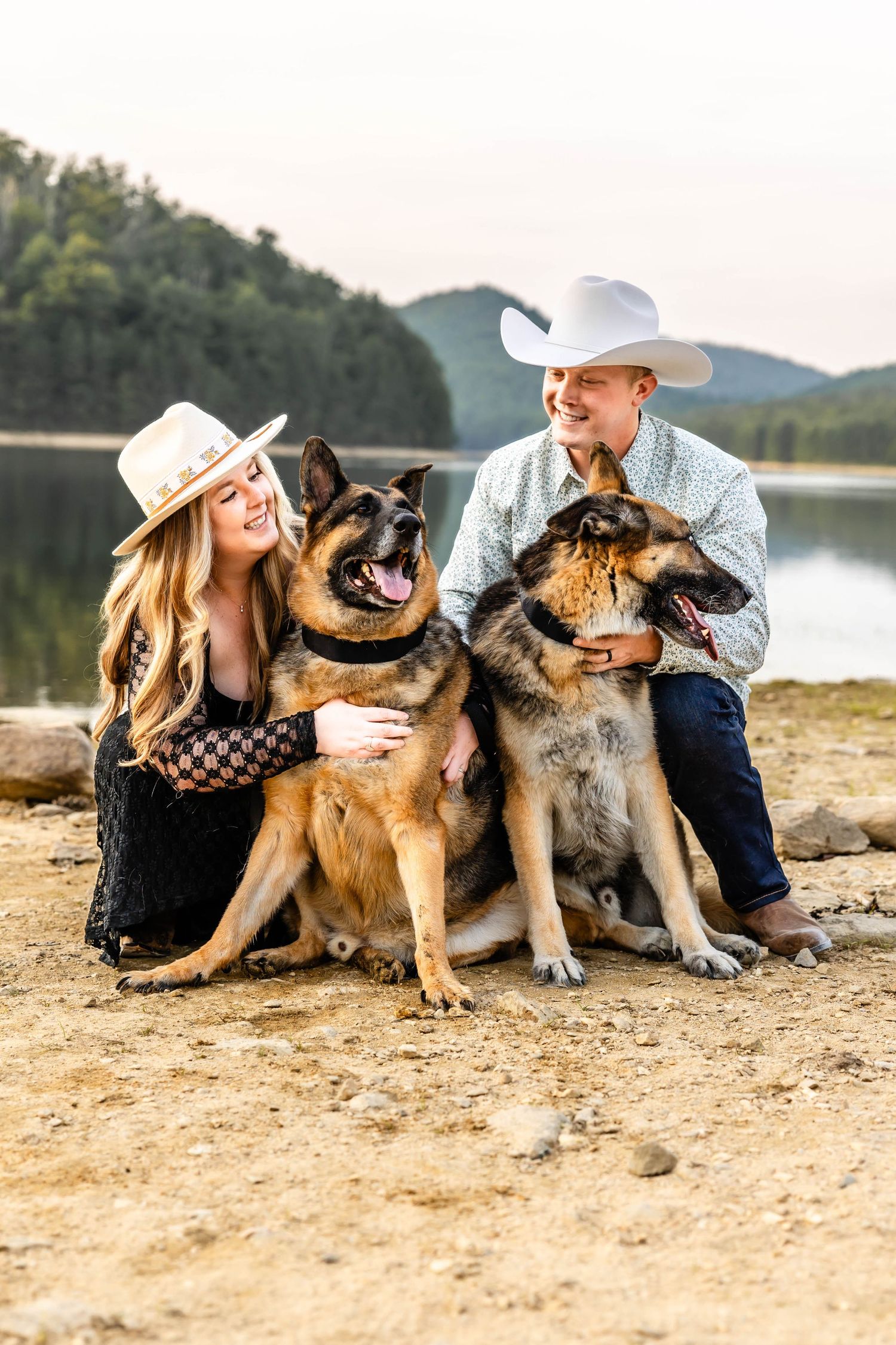 Two happy German Shepherds sit with their owners by a lake, all wearing cowboy hats.