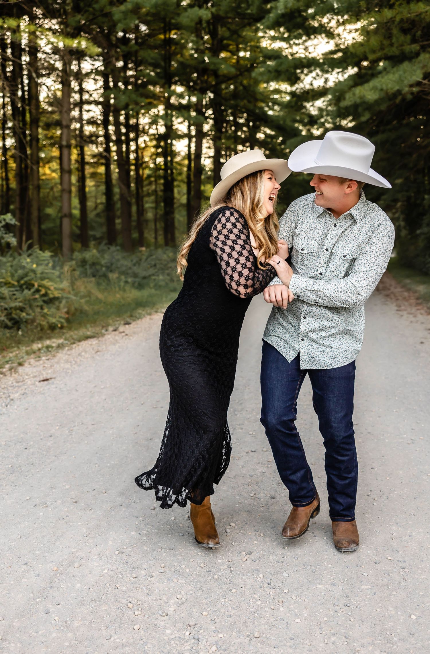 A playful couple in western wear dancing and laughing together on a dirt path surrounded by pine trees.
