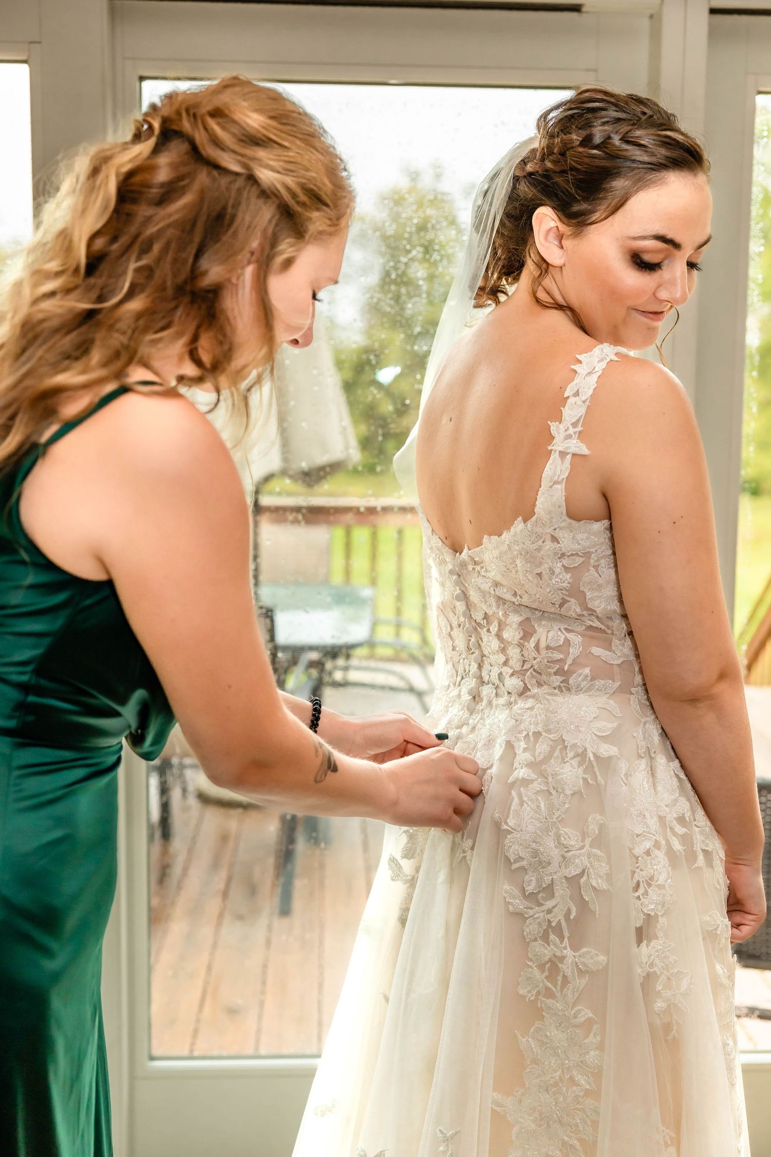 A bridesmaid in emerald green helps adjust the back of a lace wedding gown.