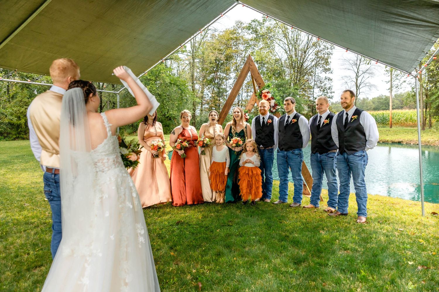 Wedding party poses under outdoor tent with bridesmaids in coral dresses and groomsmen in casual attire.