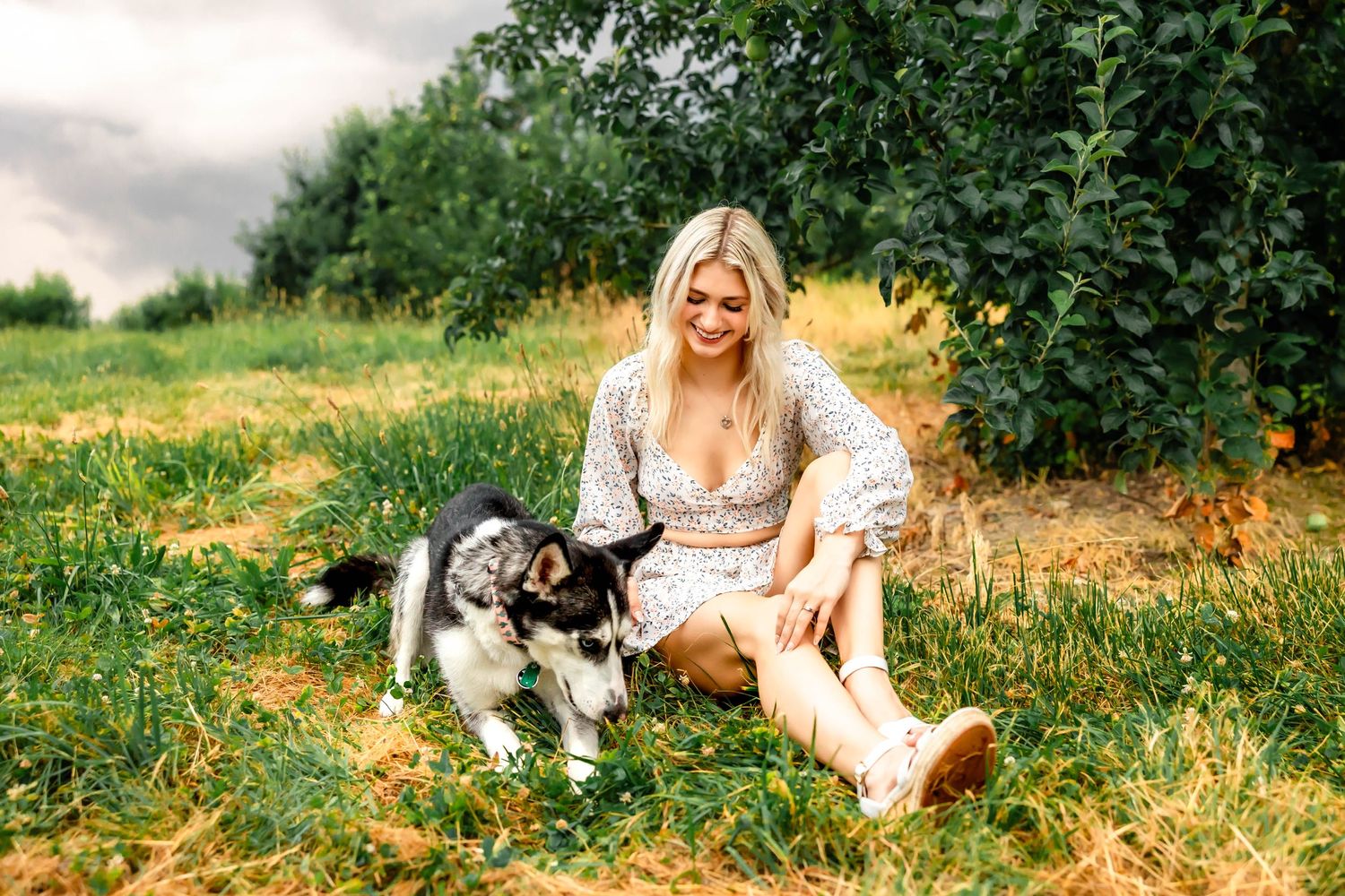 Woman in white dress sitting outdoors with playful Husky dog in natural setting.