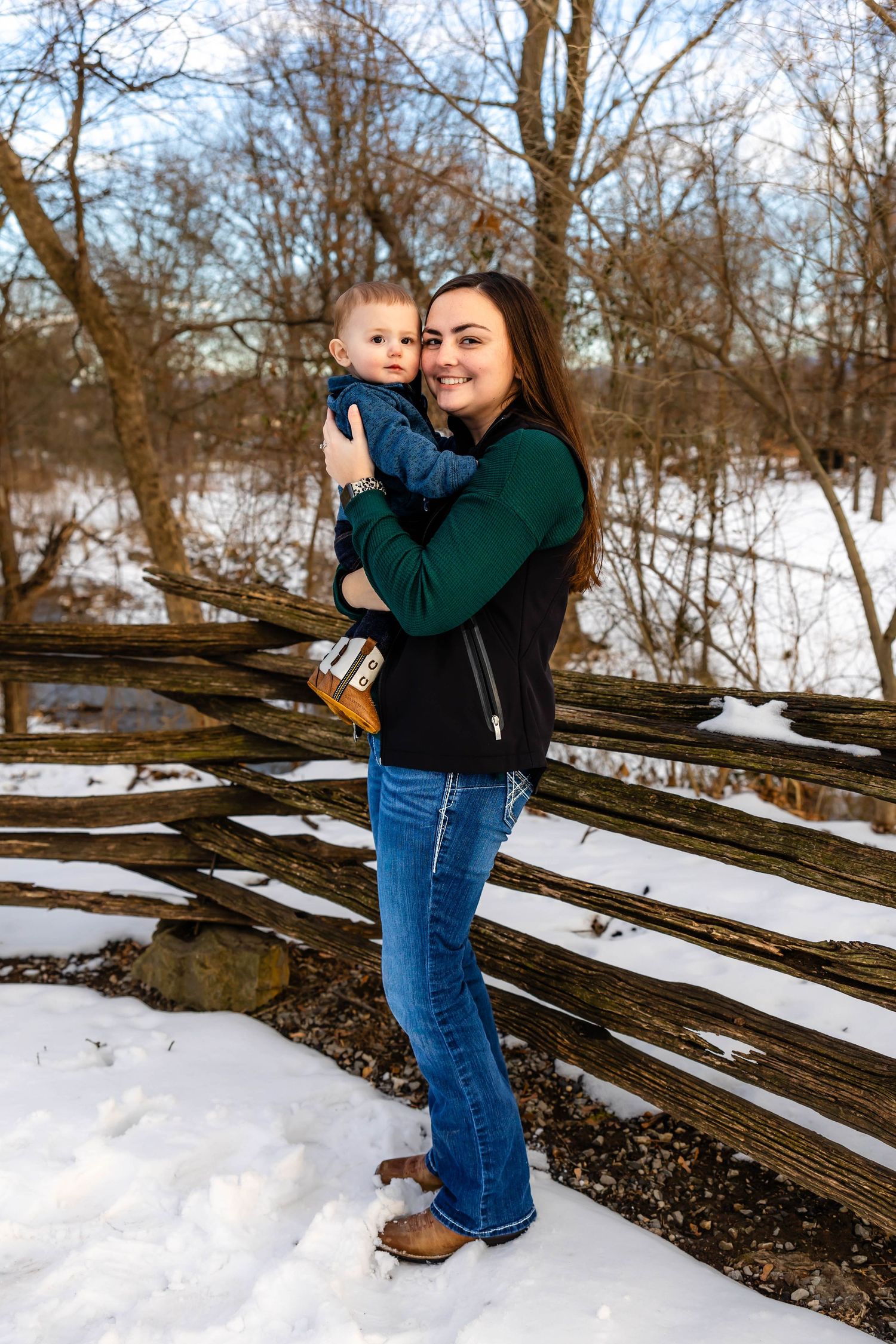 Mother holds young child against snowy landscape with wooden fence backdrop.