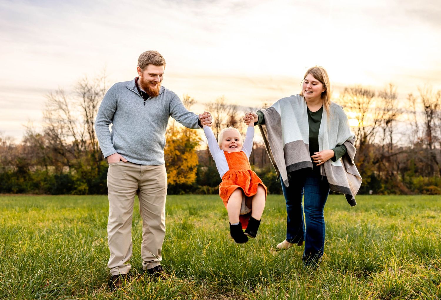 Family swinging small child in orange dress between them in grassy field at sunset.