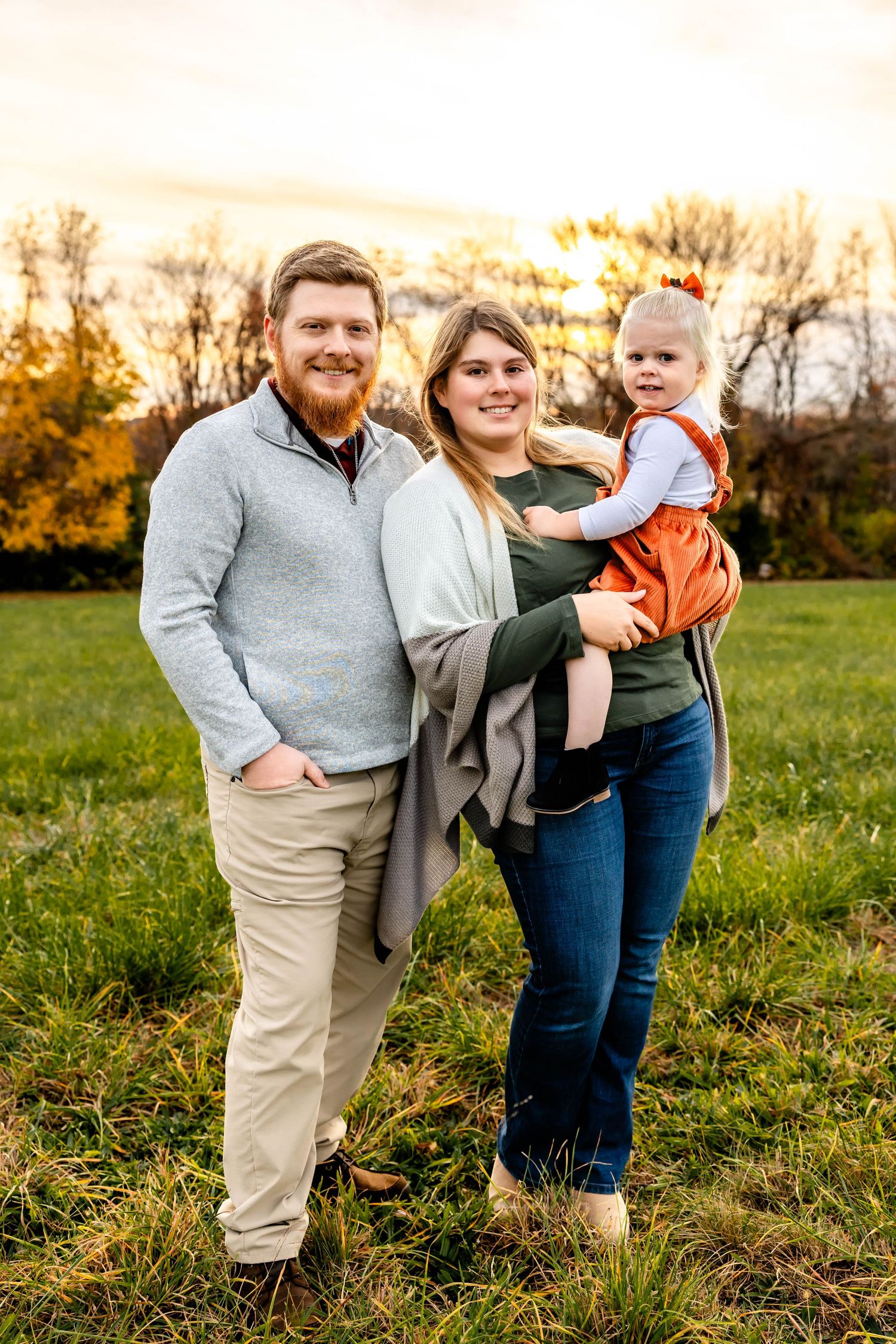 Family poses for portrait in casual fall attire against sunset backdrop.