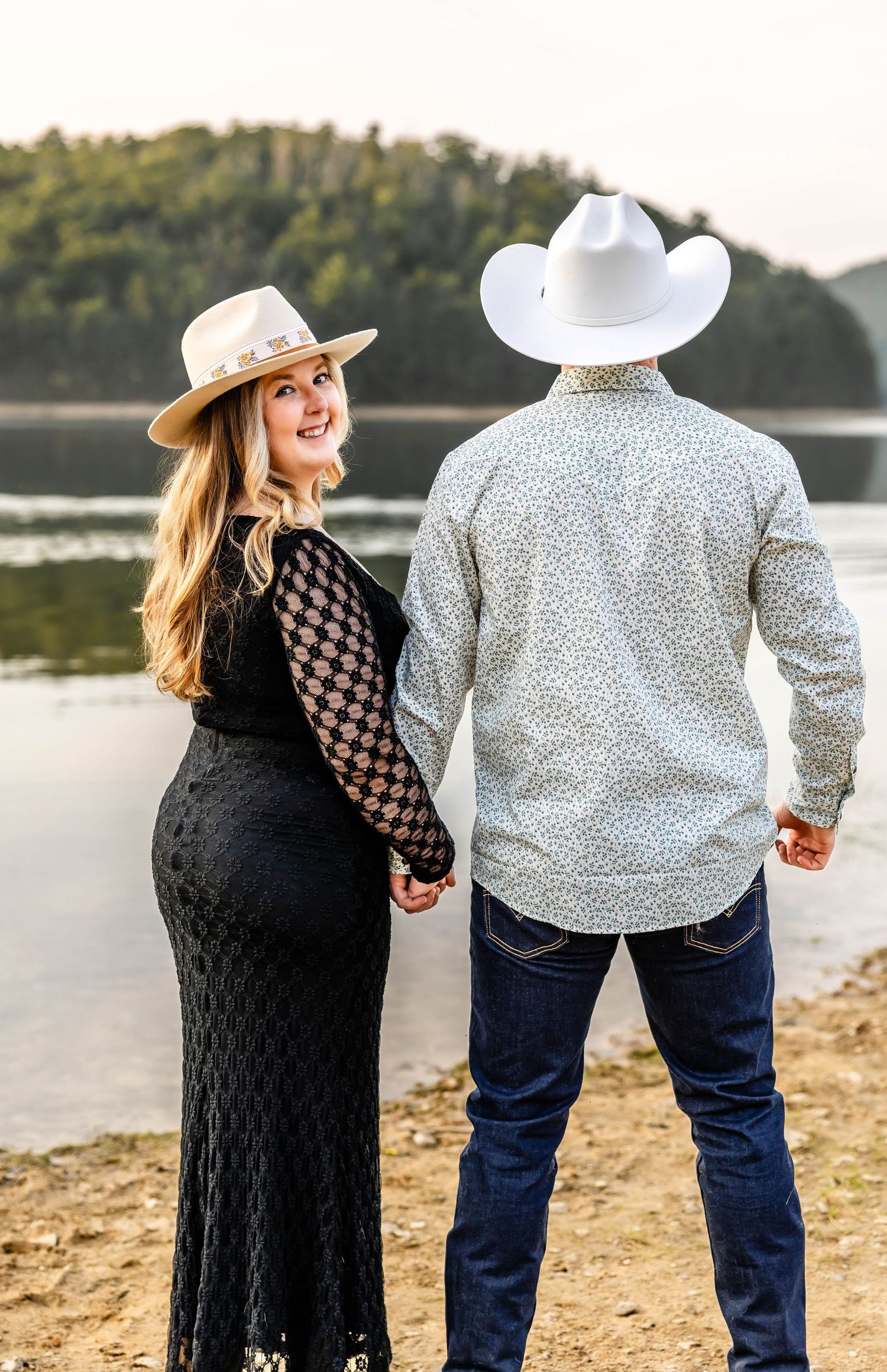 Woman in black dress and cowboy hat holds hands with partner while looking back by the lake.