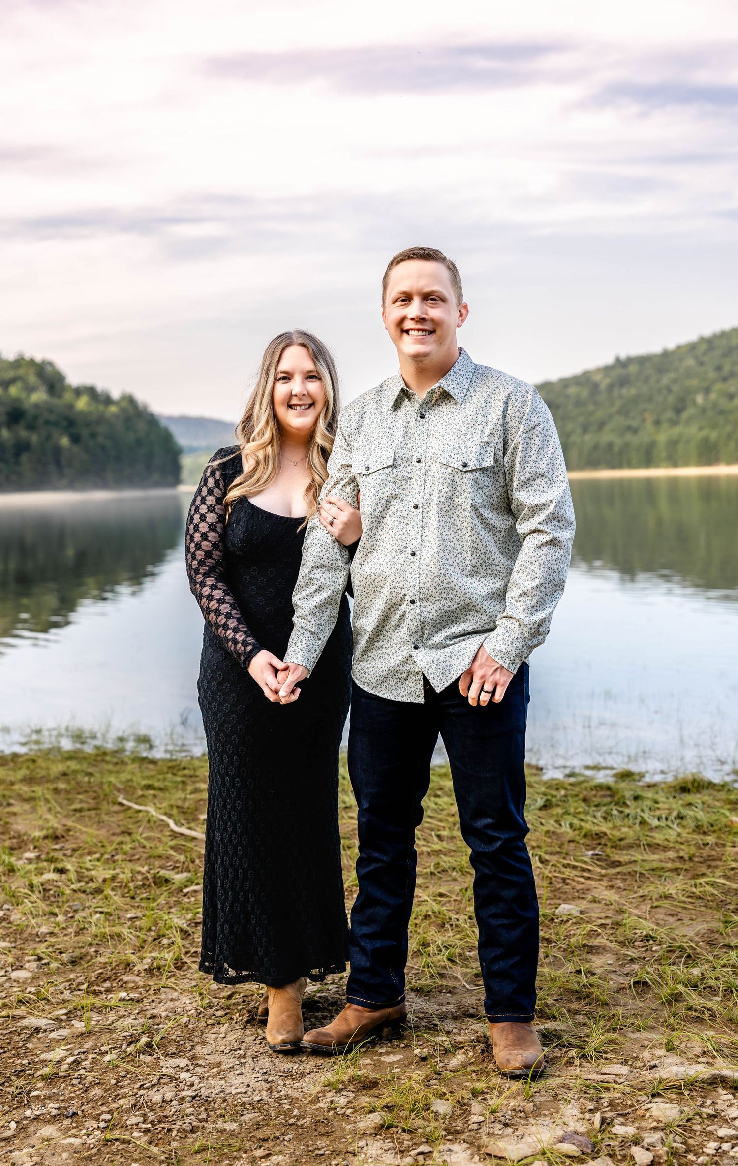 Couple poses by serene lake surrounded by mountains during engagement photoshoot.