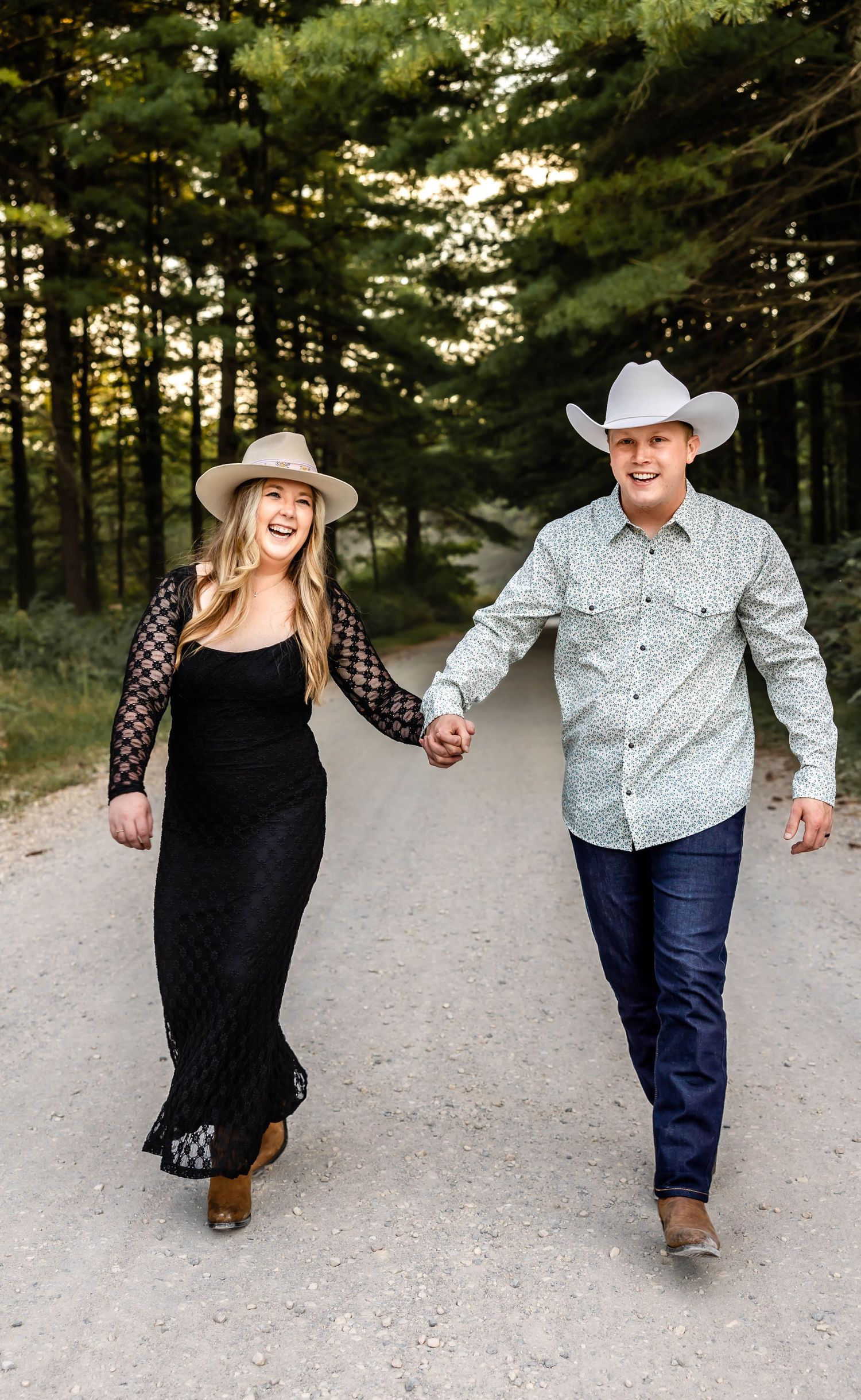 A couple laughing while walking together on a forest trail wearing western-style clothing.
