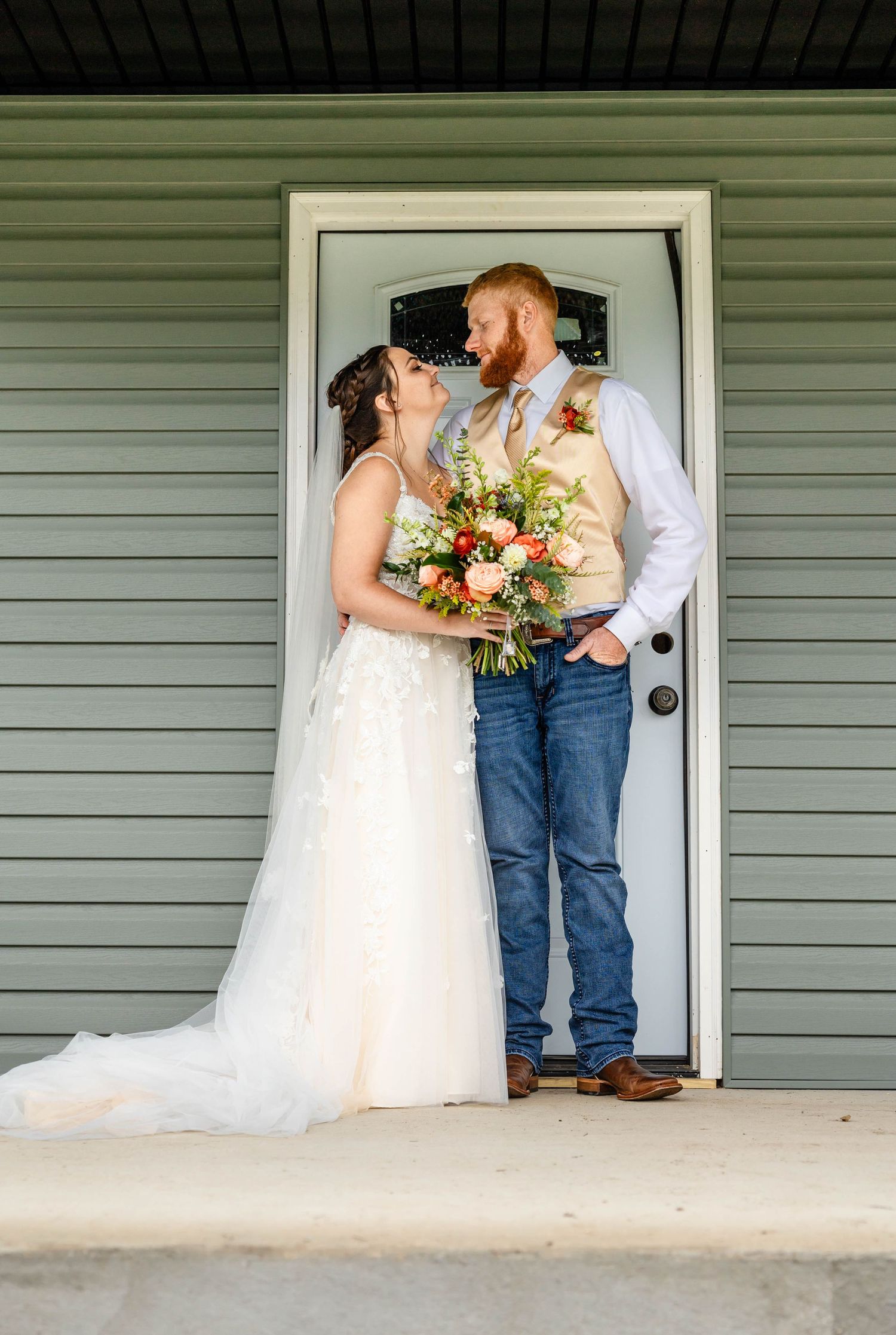 A rustic wedding couple poses in front of a sage green door, with the bride holding a coral and peach bouquet.