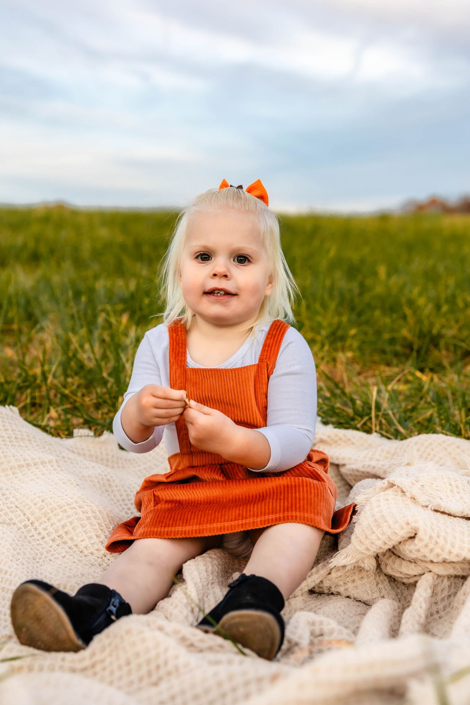 Small child in orange pinafore dress sitting on knit blanket outdoors.