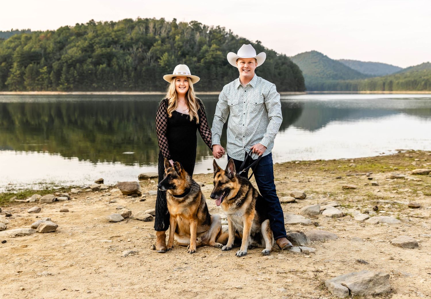A couple in western wear poses with their two German Shepherd dogs by a serene lake.