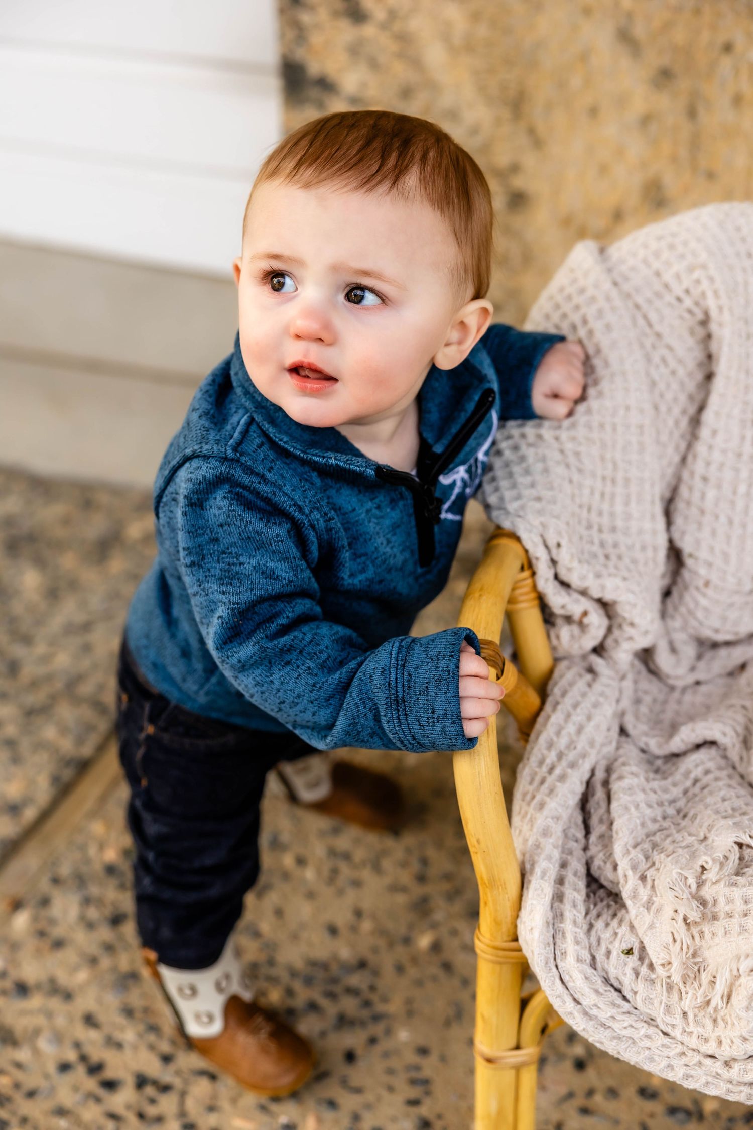 Toddler in denim and blue sweater leaning on yellow chair with knit blanket.