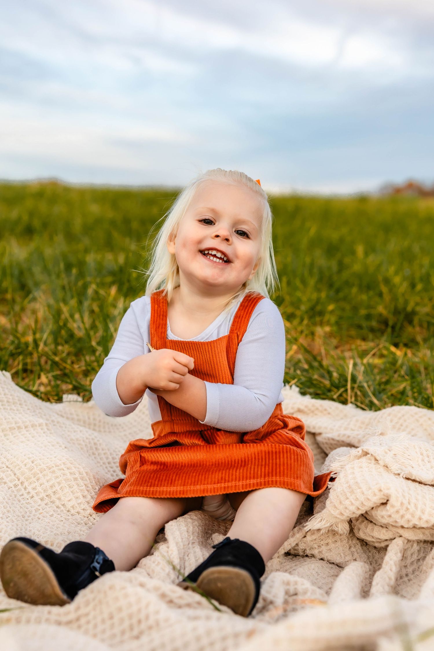 Happy child wearing orange dress and white shirt sitting on blanket outdoors.