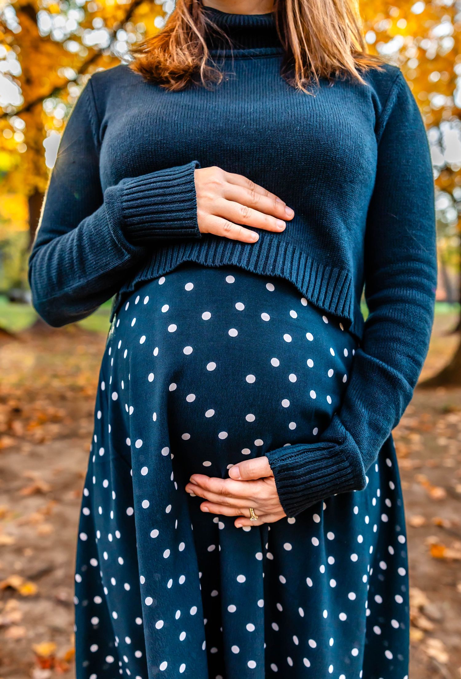 Close up of pregnant belly in navy polka dot dress and sweater against fall backdrop.