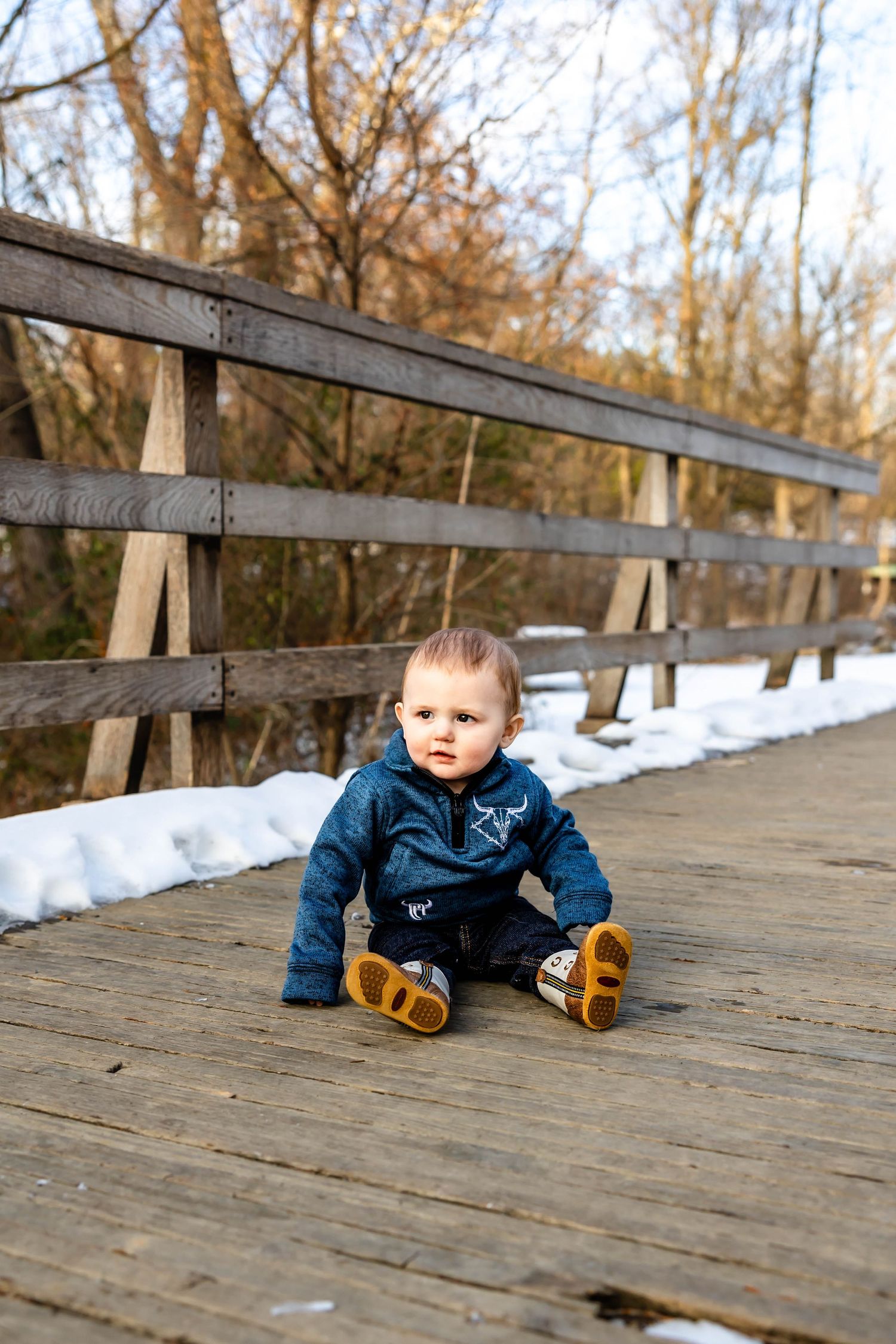 Baby wearing navy blue outfit sits on wooden bridge during winter.