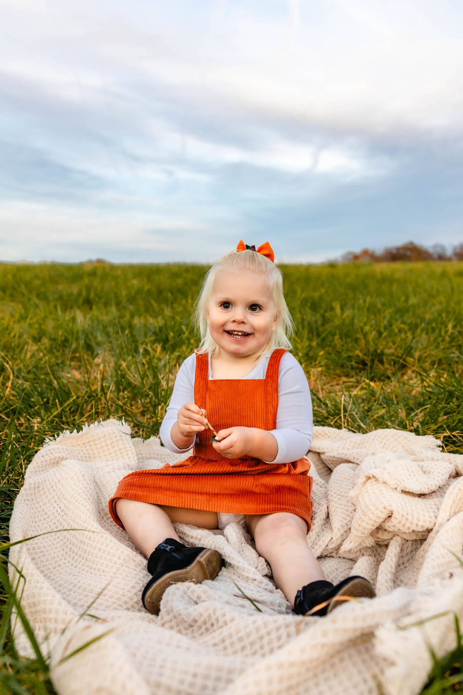 A toddler in orange overalls and bow sits on a cream blanket outdoors with a bright smile.