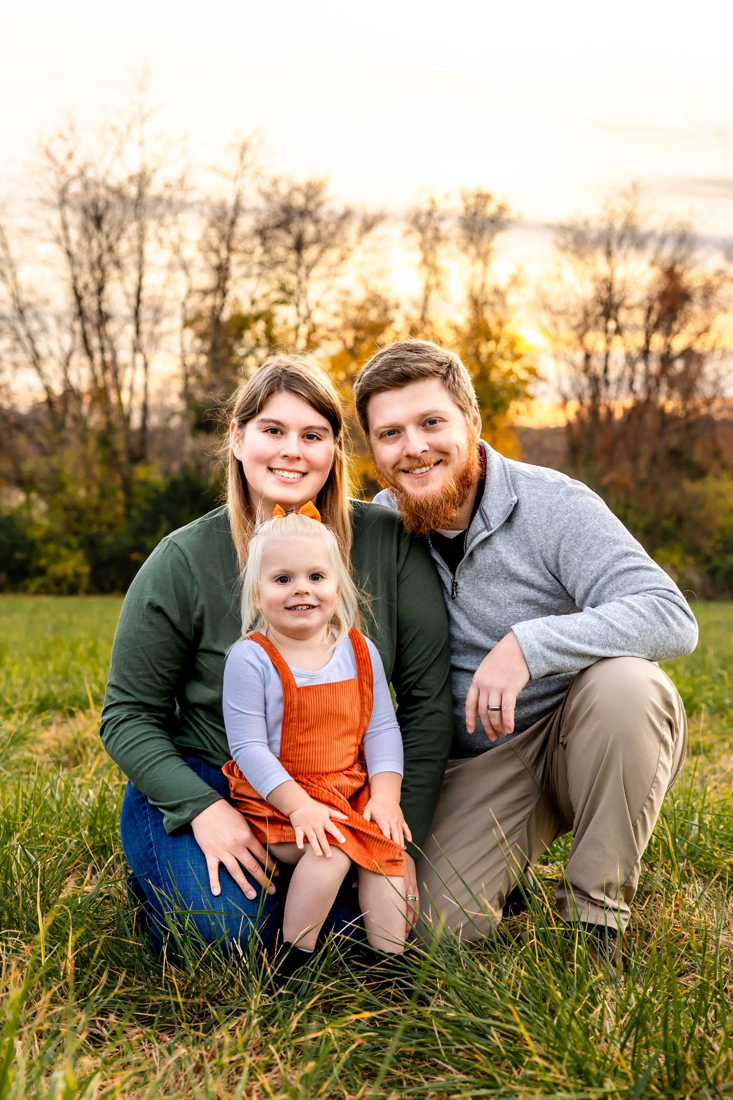 Family portrait outdoors in field during golden hour with autumn trees in background.