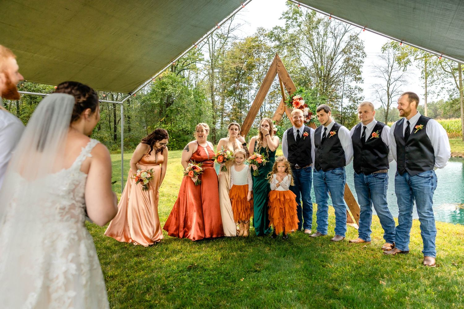 Wedding group stands in formation under white tent with bridesmaids wearing orange ombre dresses.