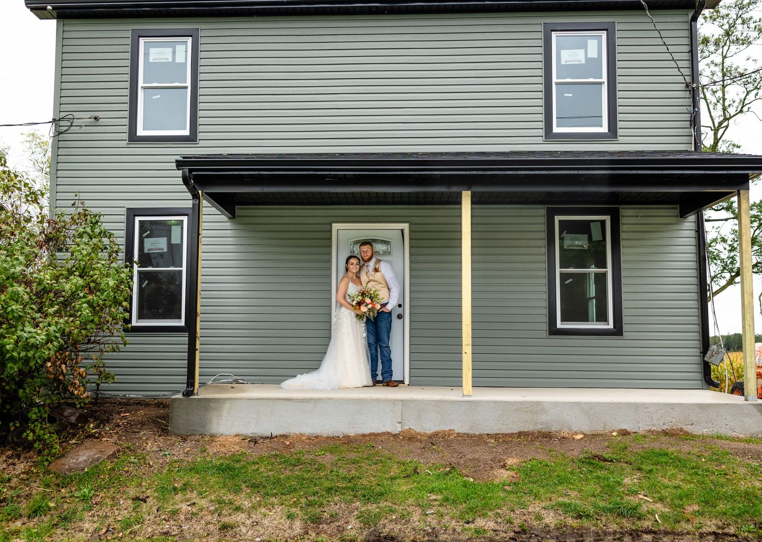 A couple poses on the front porch of a sage green two-story house.