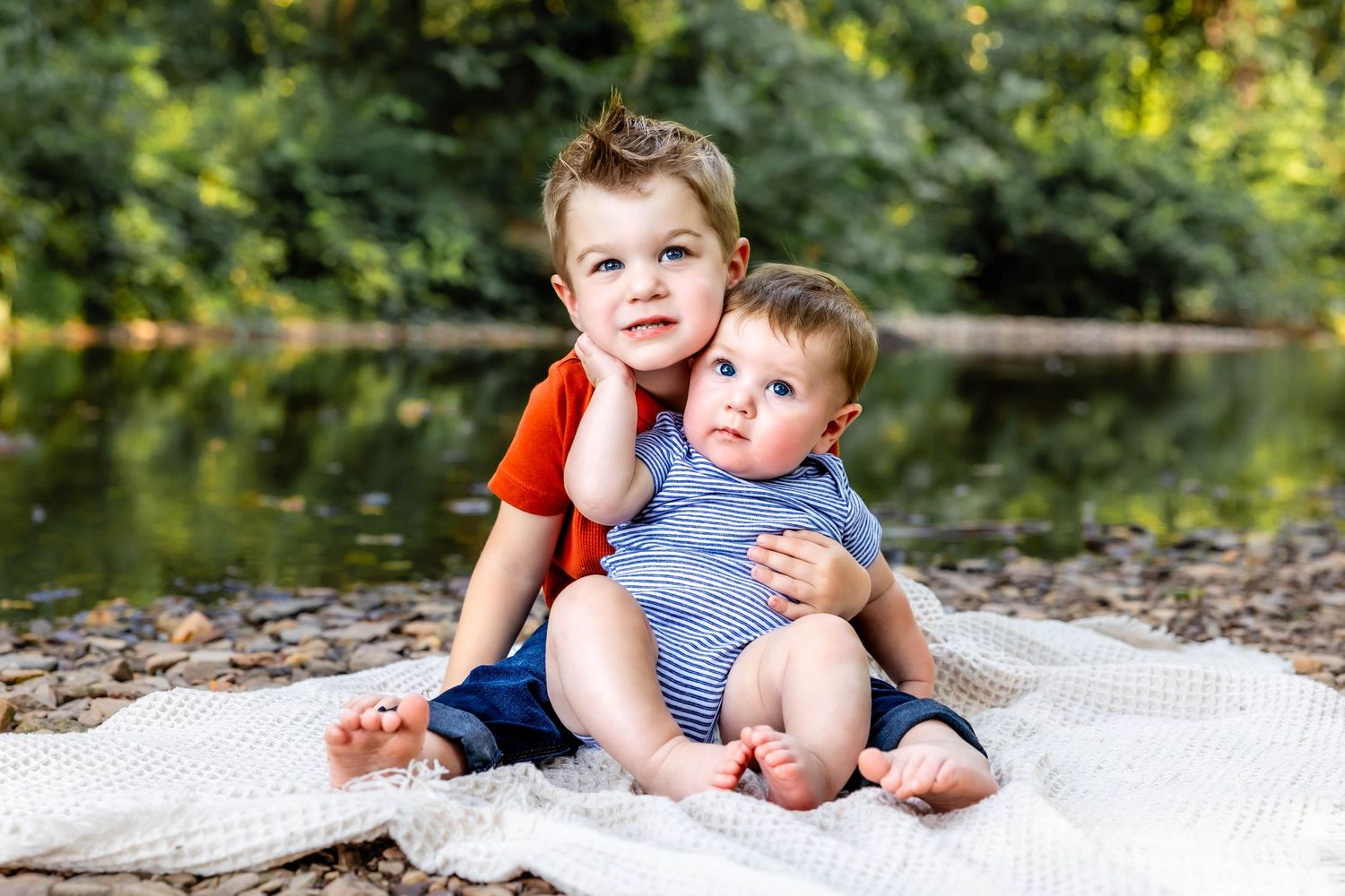 Two young siblings sitting together on a blanket by a peaceful pond.