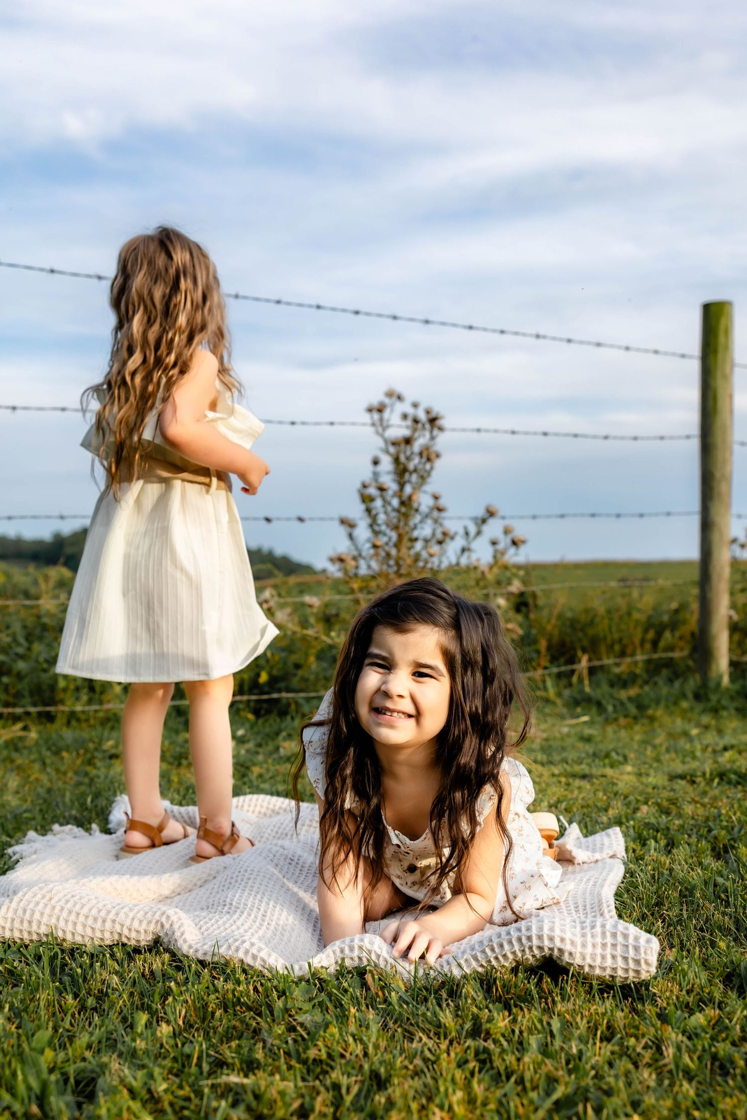 Two children in white dresses enjoying a sunny day on a blanket near a barbed wire fence in a grassy field.
