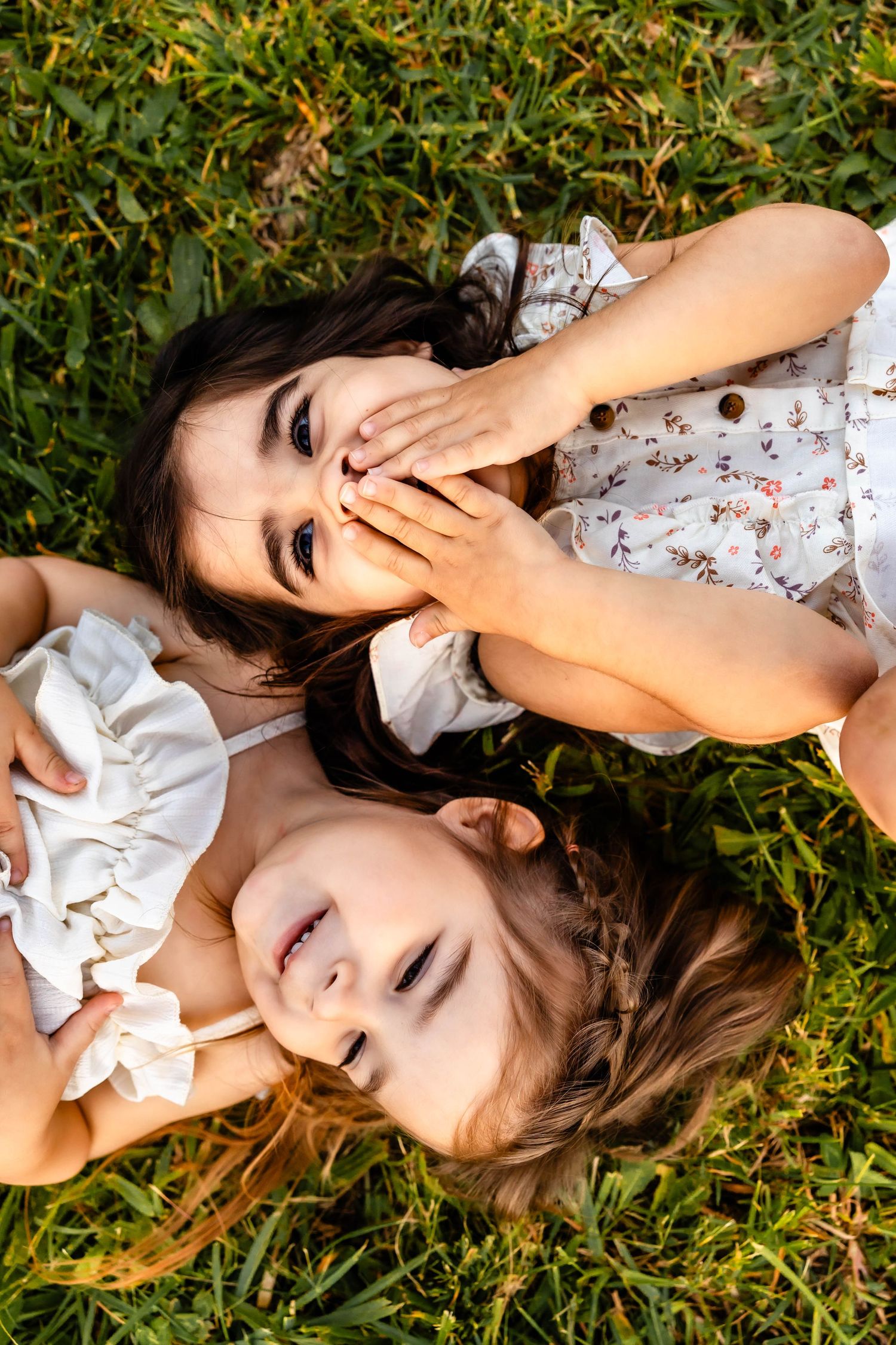 Two people lying head-to-head in grass, giggling and covering their smiles with their hands.